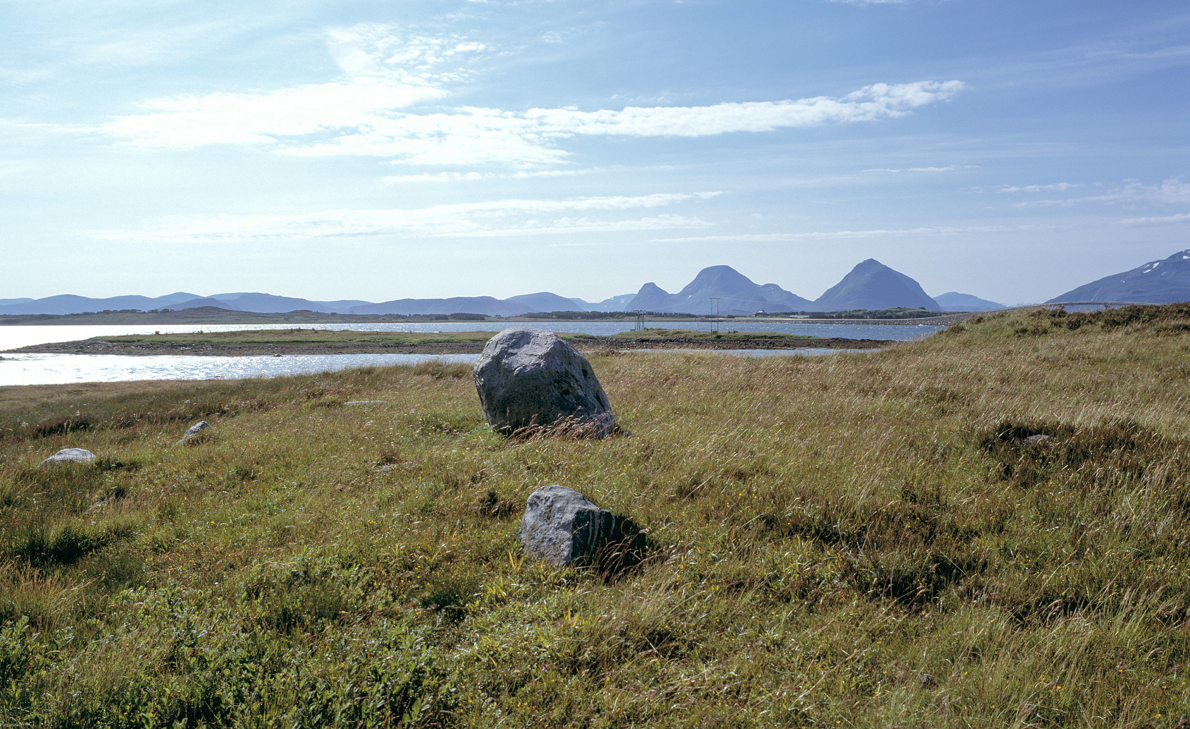 Dypdykk i våre arkeologiske kulturminner 1 Steinete gressletter nær et rolig vann med fjell i det fjerne under en delvis overskyet himmel.