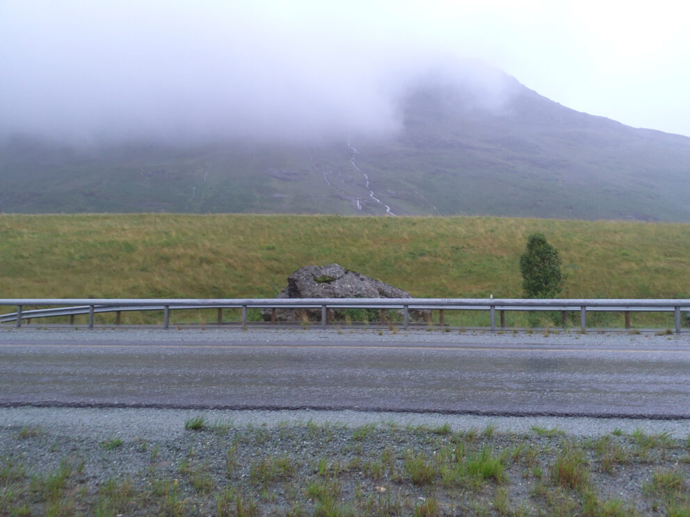 Dypdykk i våre arkeologiske kulturminner 6 En våt vei med et metallrekkverk, en gressbakke, en stor stein, et lite tre og et tåkete fjell i bakgrunnen som delvis er dekket av skyer.