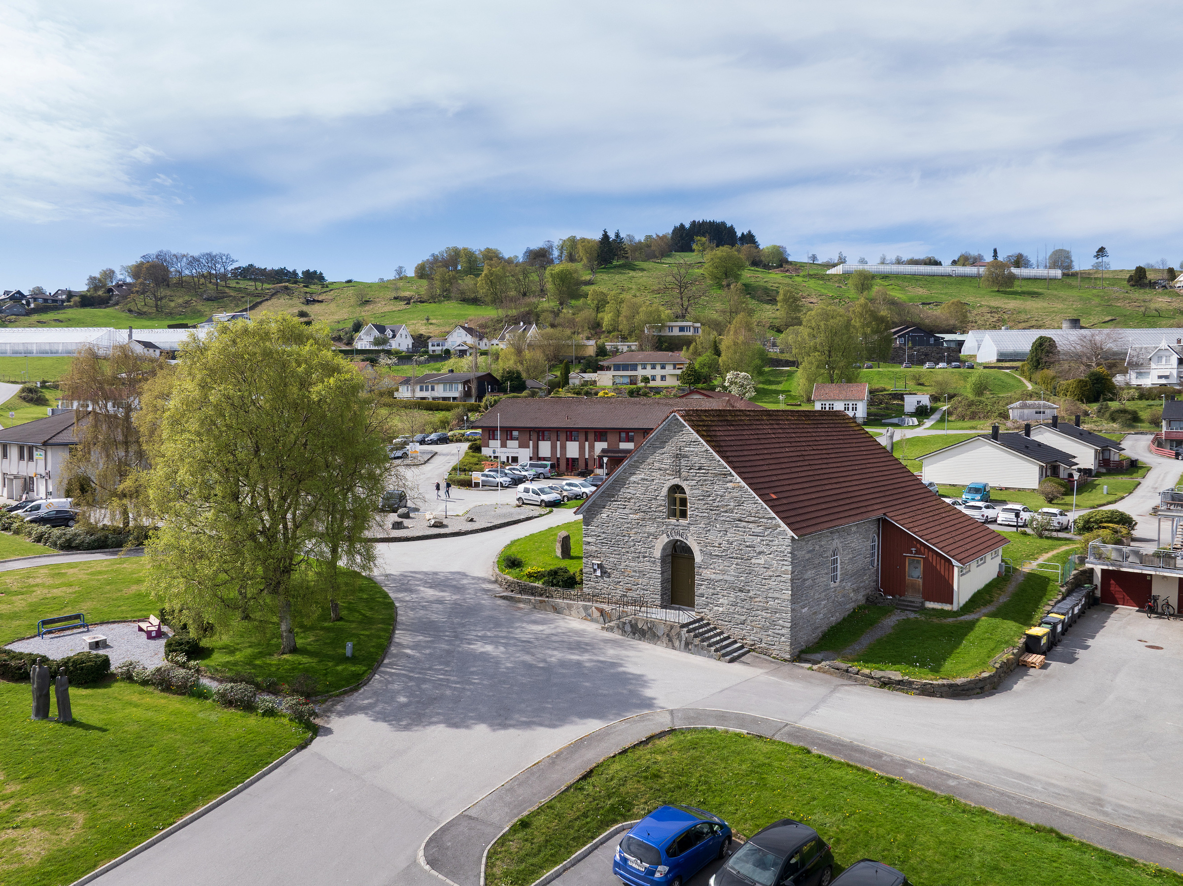 En liten steinkirke med rødt tak står i en landlig landsby omgitt av grønne åser, hus, parkerte biler og trær under en delvis overskyet himmel.