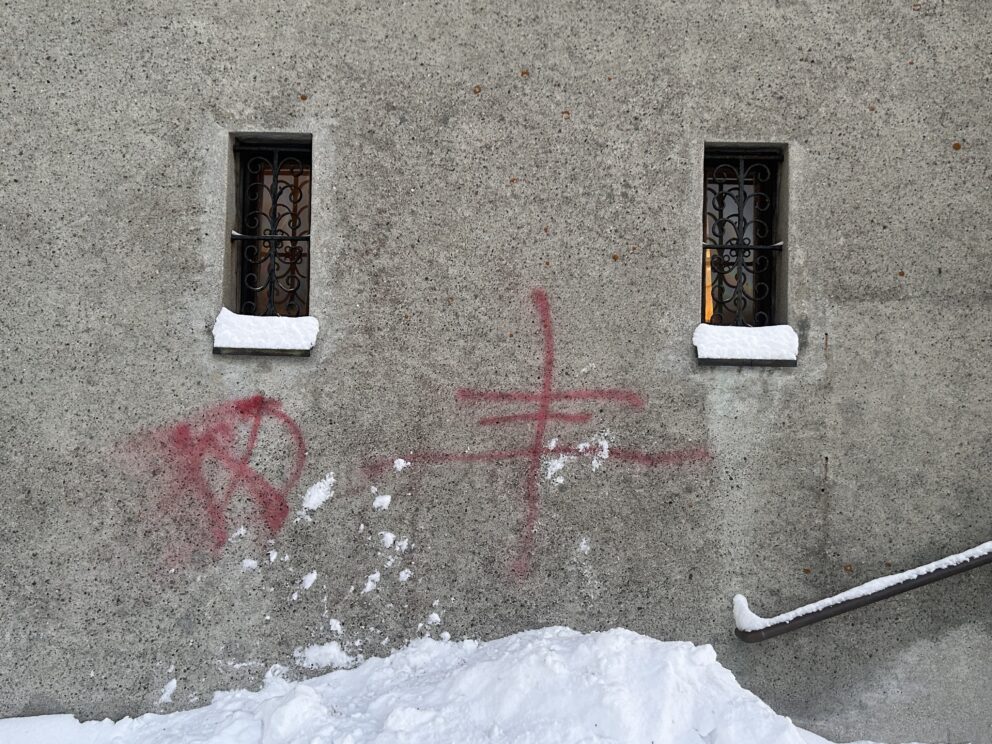 Graffiti i rød spraymaling på en betongvegg med to sprossede vinduer, snø på avsatser og bakken, og et metallrekkverk synlig nederst til høyre.