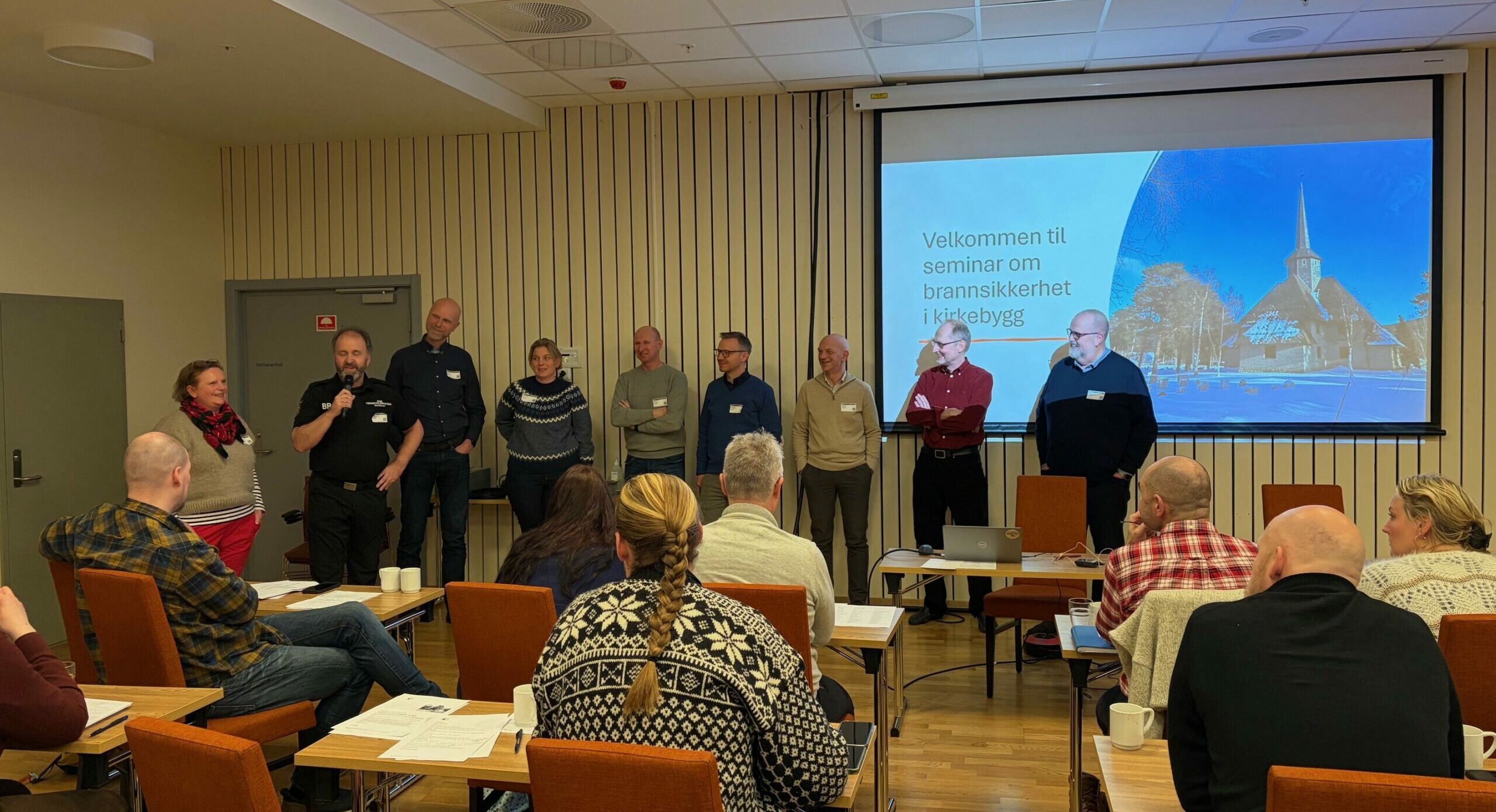 En gruppe mennesker står foran i et konferanserom og henvender seg til de sittende deltakerne. Bak dem projiseres et presentasjonsbilde om brannsikkerhet.
