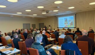En gruppe mennesker sitter i et konferanserom foran en prosjektorskjerm og lytter til en presentasjon.