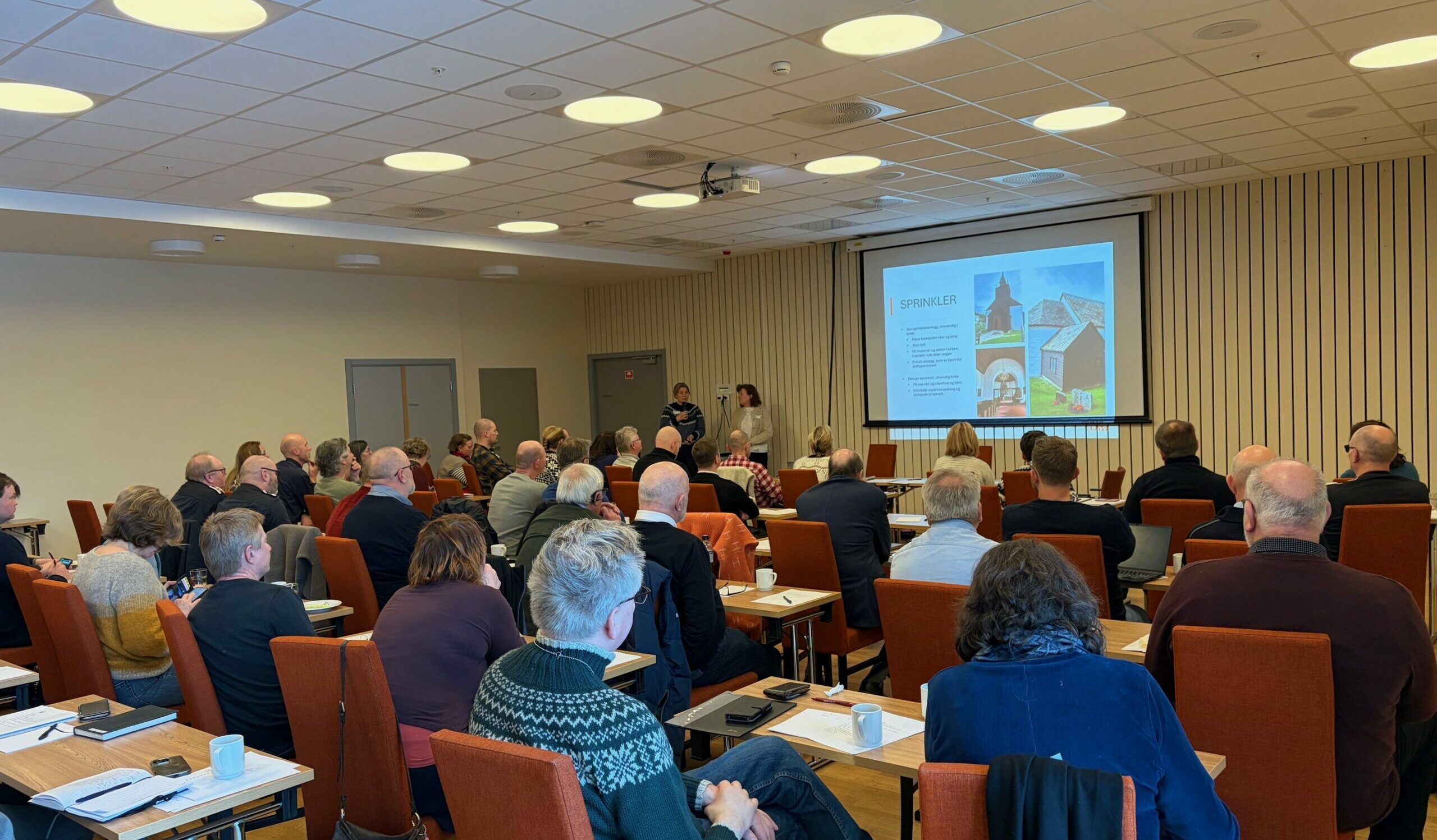 En gruppe mennesker sitter i et konferanserom foran en prosjektorskjerm og lytter til en presentasjon.