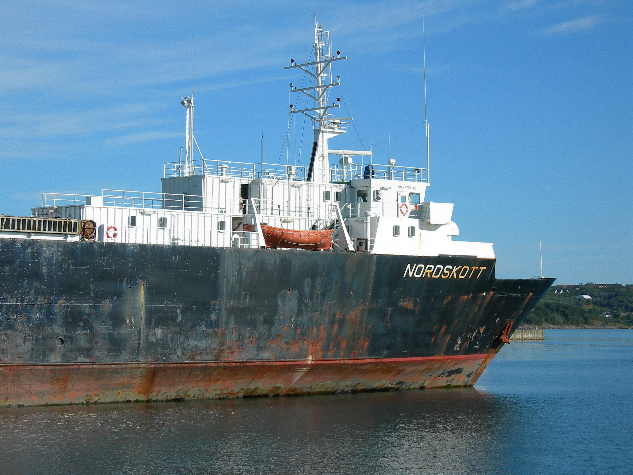 Et stort lasteskip ved navn NORDSKOTT ligger til kai i en havn, med synlig rust langs det svarte skroget og en blå himmel i bakgrunnen.