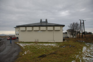 En beige bygning med flatt tak, små vinduer og to skorsteiner står under en overskyet himmel, omgitt av gressflekker, snø og en grusvei.