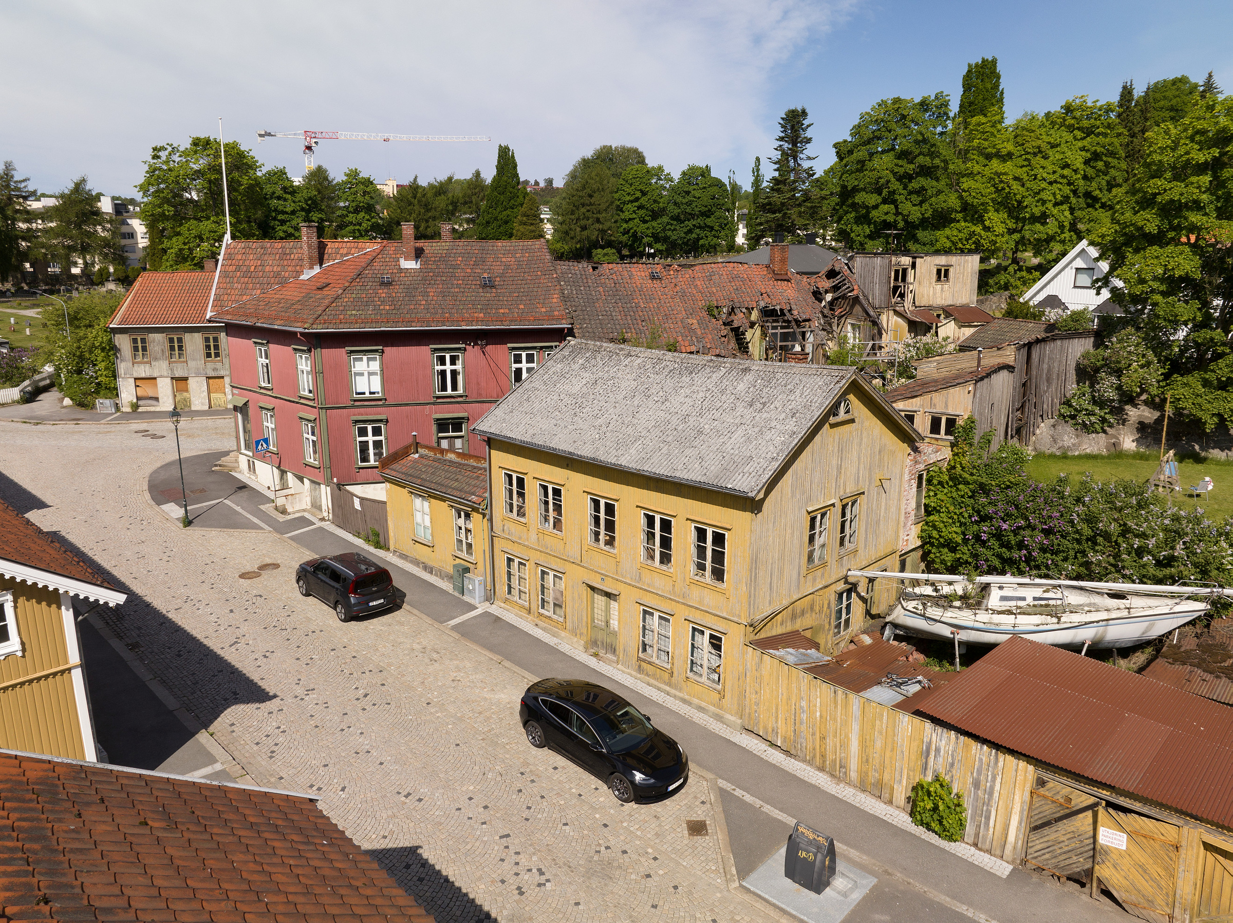 En gate med parkerte biler ved siden av gamle trebygninger, den ene gul og forvitret, omgitt av trær og grøntområder under en delvis overskyet himmel.