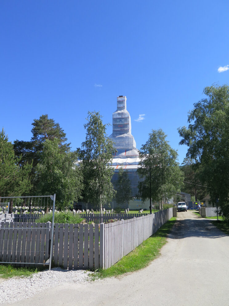 En høy bygning under oppføring eller renovering er dekket av stillaser og hvite plater, omgitt av trær, et tregjerde og en klar, blå himmel.