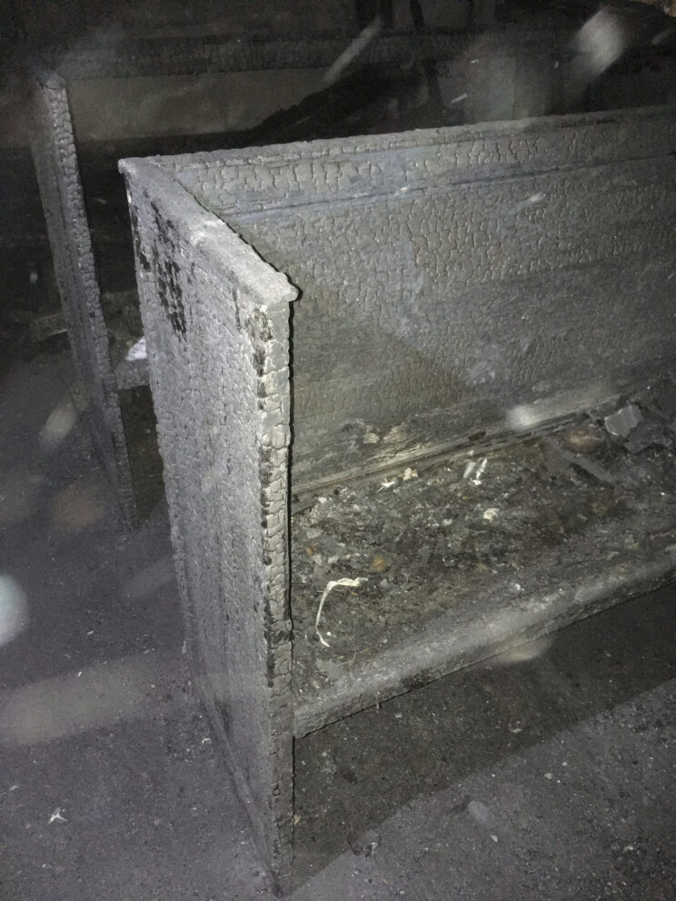 En kirkebenk som er kraftig forkullet og dekket av sot fra en brann nylig, står i et mørkt, søppelstrødd område.