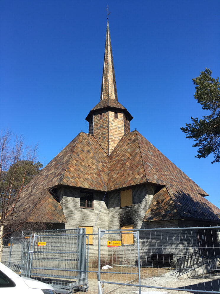 En kirke med bratt sponkledd tak og høyt spir, omgitt av et metallgjerde og bordkledde vinduer, under en klarblå himmel.