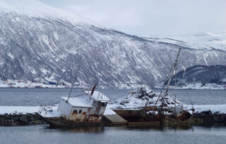To rustne, delvis sunkne båter - et stemningsfullt skipsfunn - hviler nær en snødekt strandlinje med fjell og vann i bakgrunnen.