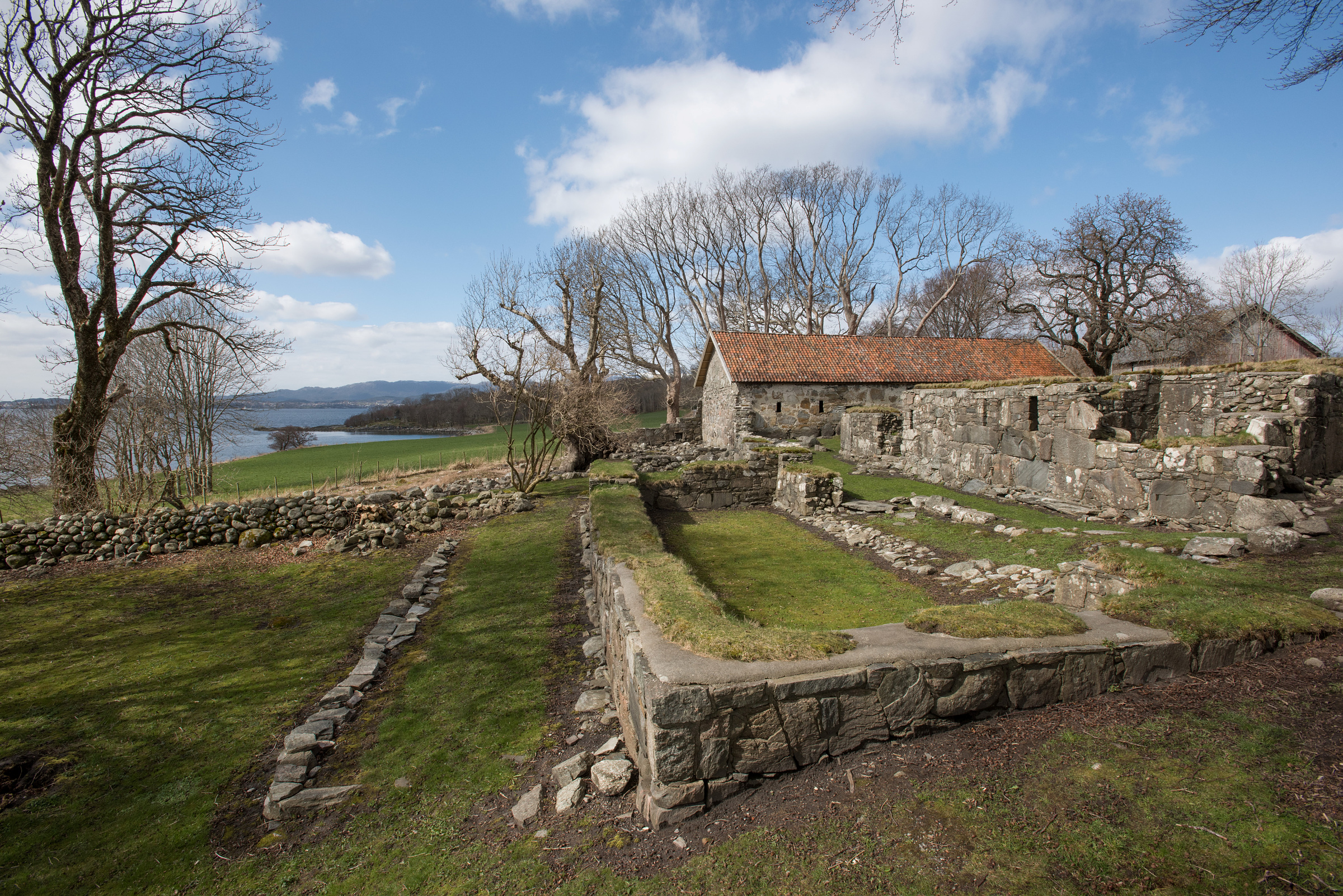 Ruiner av en gammel steinbygning med delvis intakt tak, omgitt av gress, trær og en steinmur, med utsikt over en vannflate under en blå himmel med skyer.