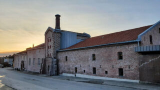 En stor industribygning i teglstein med rødt tegltak og høy skorstein, fotografert i solnedgang fra den andre siden av en stille gate.
