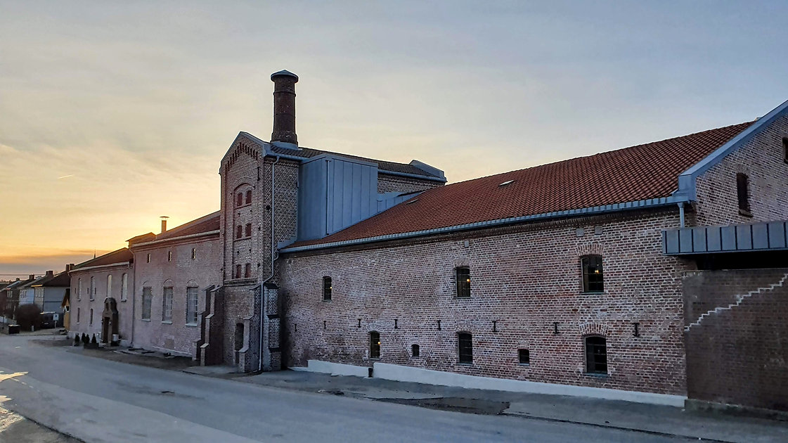 En stor industribygning i teglstein med rødt tegltak og høy skorstein, fotografert i solnedgang fra den andre siden av en stille gate.