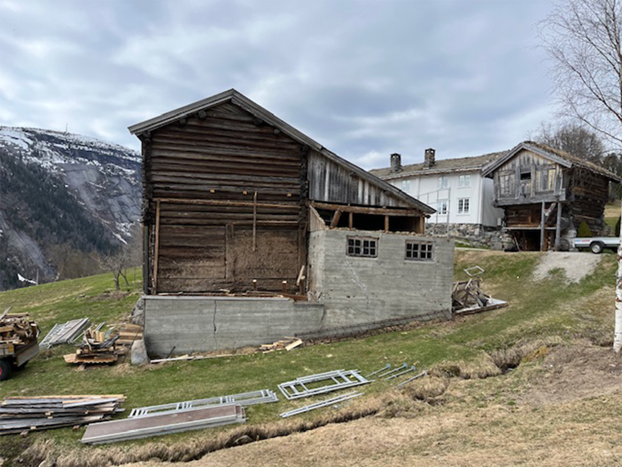 Et gammelt trebygg med betongfundament står på skrånende gress i nærheten av to gamle hus, med fjell og skyet himmel i bakgrunnen. Byggematerialer ligger spredt i nærheten.