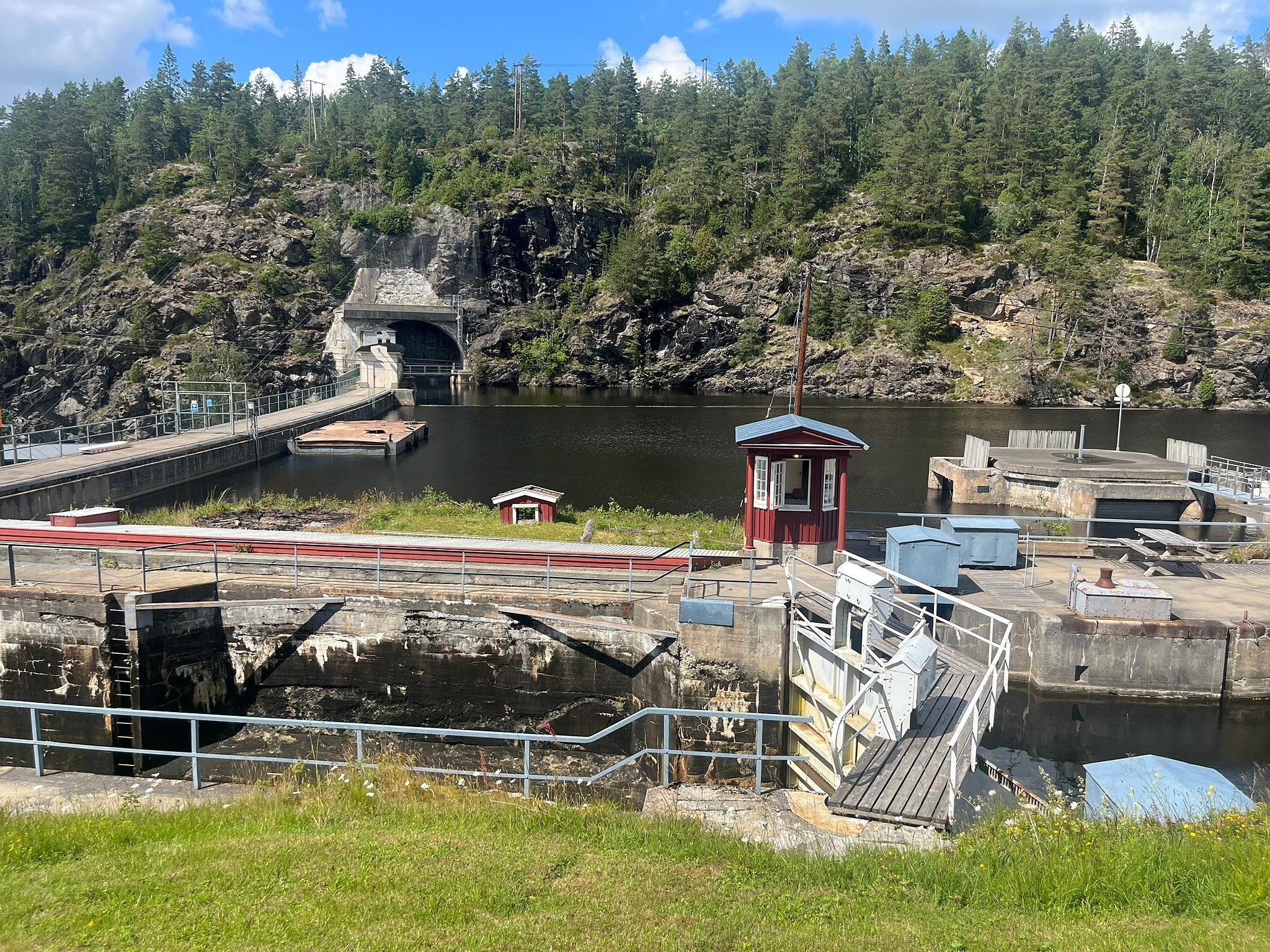 En vannkraftdam med en kanal, en rød kontrollboks, rekkverk og en tunnelinngang mot en steinete, skogkledd åsside.