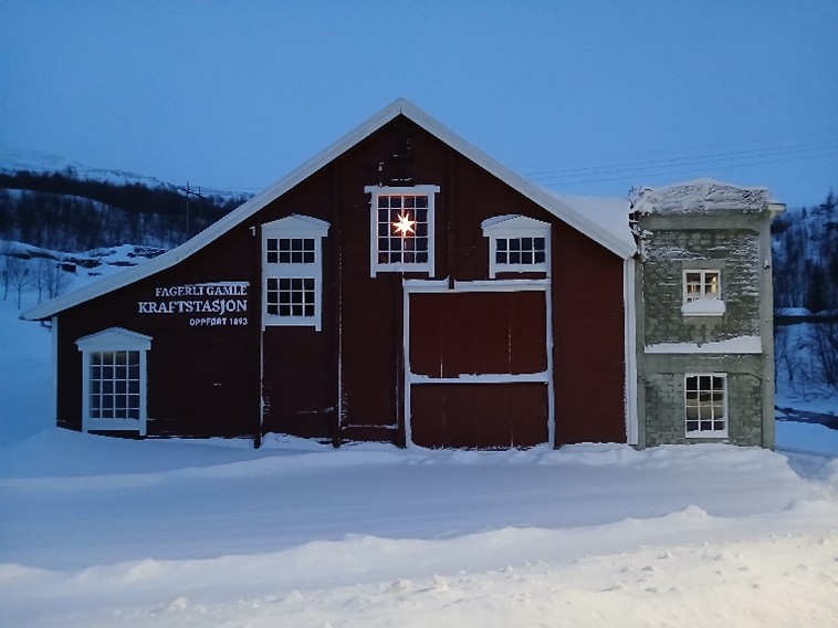 En rød bygning i tre og grå stein, merket Fagerlie Gamle Kraftstasjon Oppført 1893, står i et snødekt landskap i skumringen, med ett opplyst vindu.
