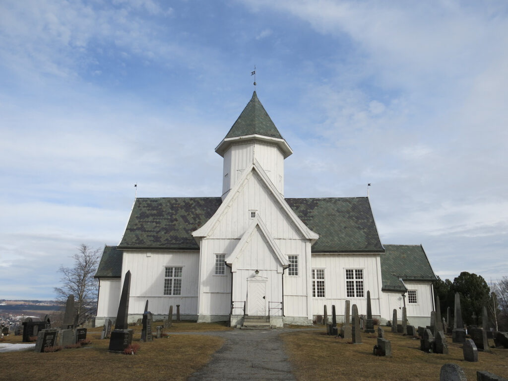 Kolbu kirke på Østre Toten. En hvit trekirke med et bratt, grønt tak står omgitt av gravsteiner under en delvis overskyet himmel.