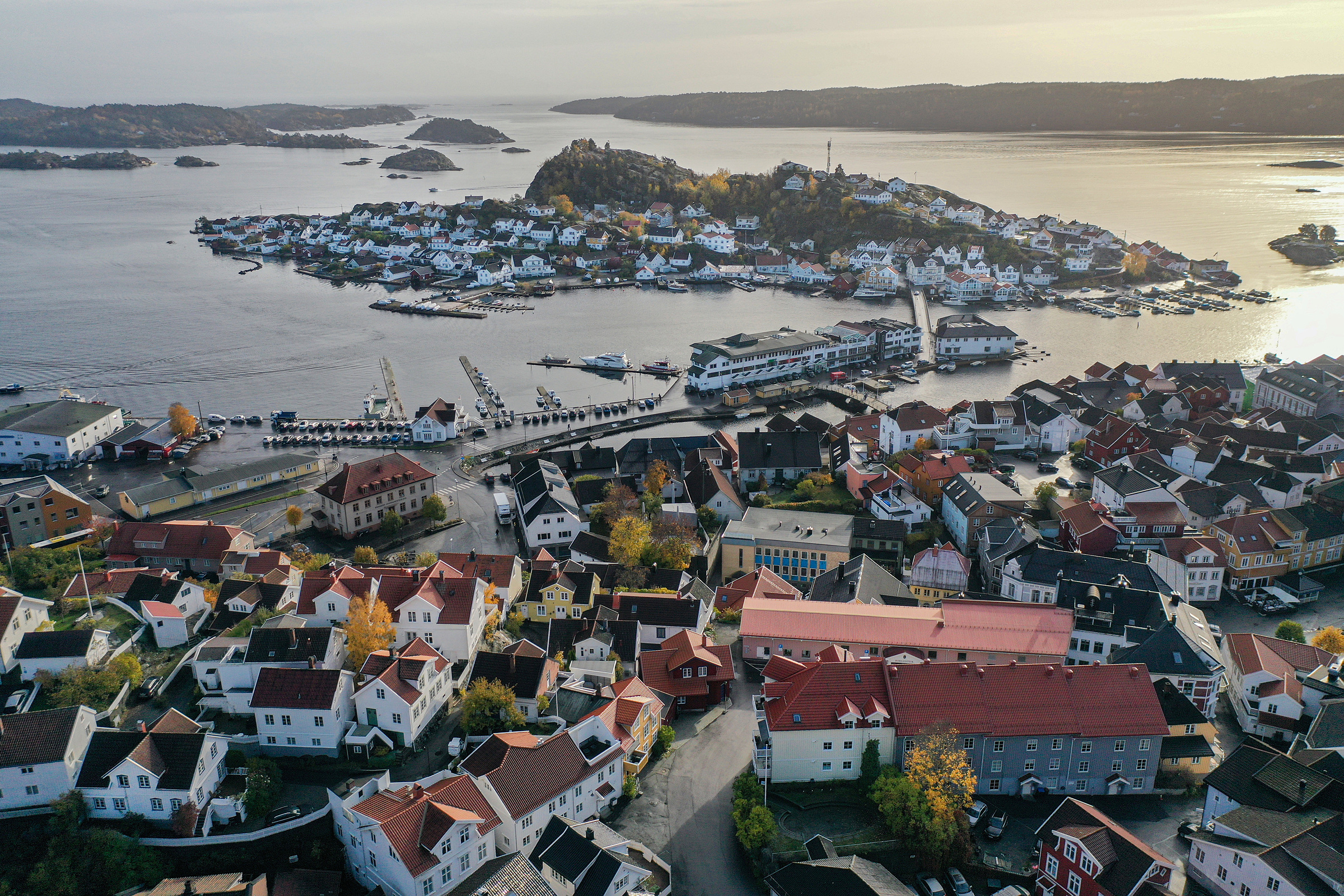 Flyfoto av en kystby med røde hus, en marina og en liten øy som er forbundet med en bro, omgitt av vann og spredte holmer.