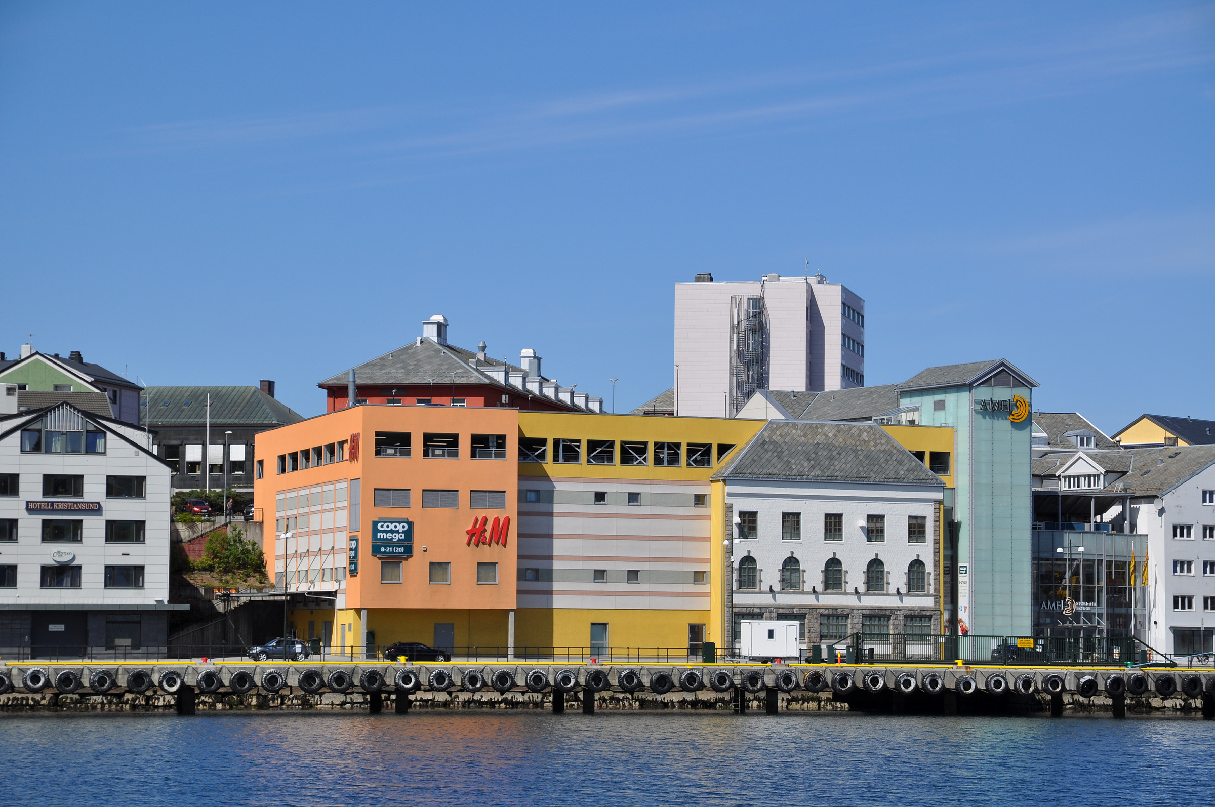 Fargerike bygninger med butikker, deriblant H&M og Coop, står langs en kai under en klarblå himmel i en kystby.