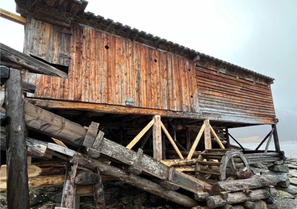 En vannmølle i tre med forvitrede planker, hevet på stylter, med vannhjul og støttebjelker, plassert i et tåkete utemiljø.