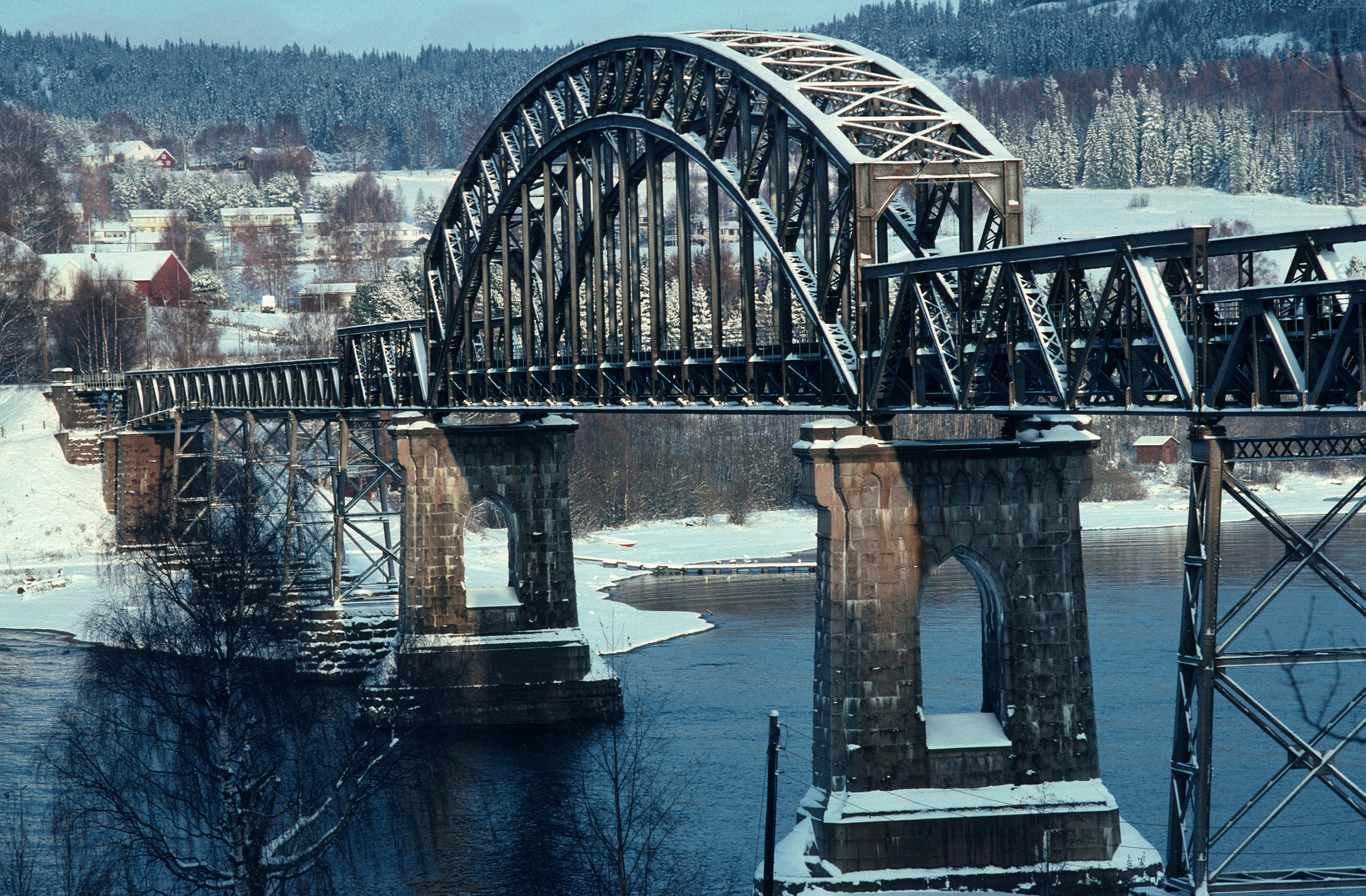 Jernbanebro i stålbue med steinpilarer over en elv i et snødekt landskap, omgitt av trær og hus i bakgrunnen.