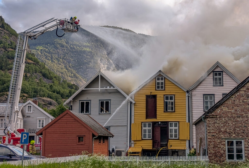 Brannmannskaper på en stigebil spruter vann på et brennende gult hus omgitt av røyk og nabohus i et fjellområde.