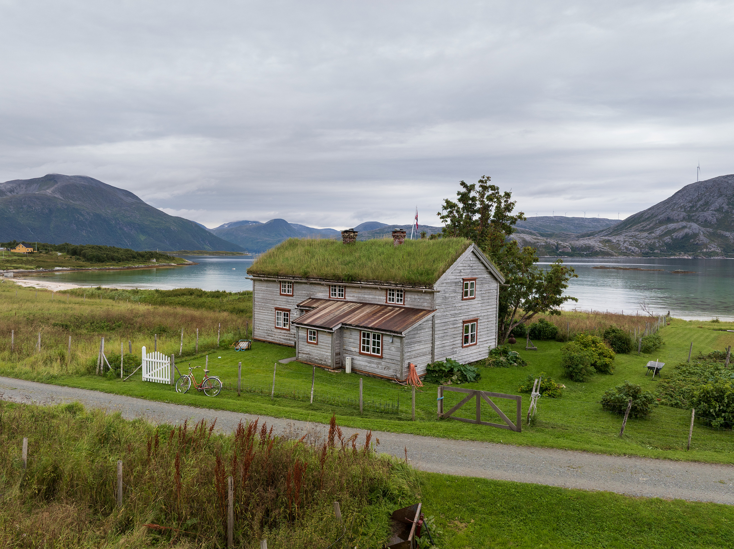 Et trehus med gresskledd tak står ved en innsjø, omgitt av grønn gressplen, fjell og overskyet himmel. Et hvitt gjerde og en sykkel står på gårdsplassen.