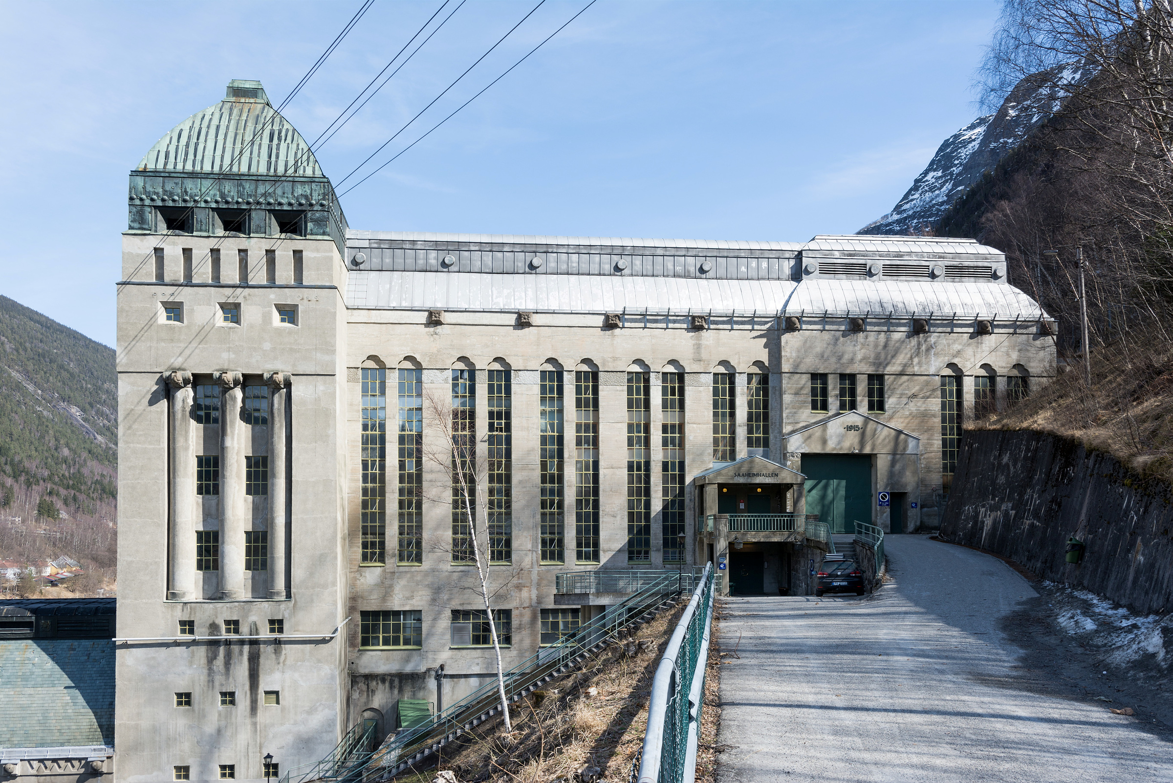 Riksantikvartimen - kulturmiljøstrategiar for bygd, by, næring og industri 3 Såheim kraftverk på Rjukan. En stor industribygning i betong med høye vinduer og et grønt metalltak, plassert mot en fjellkulisse med kraftledninger som strekker seg ut fra bygningen.