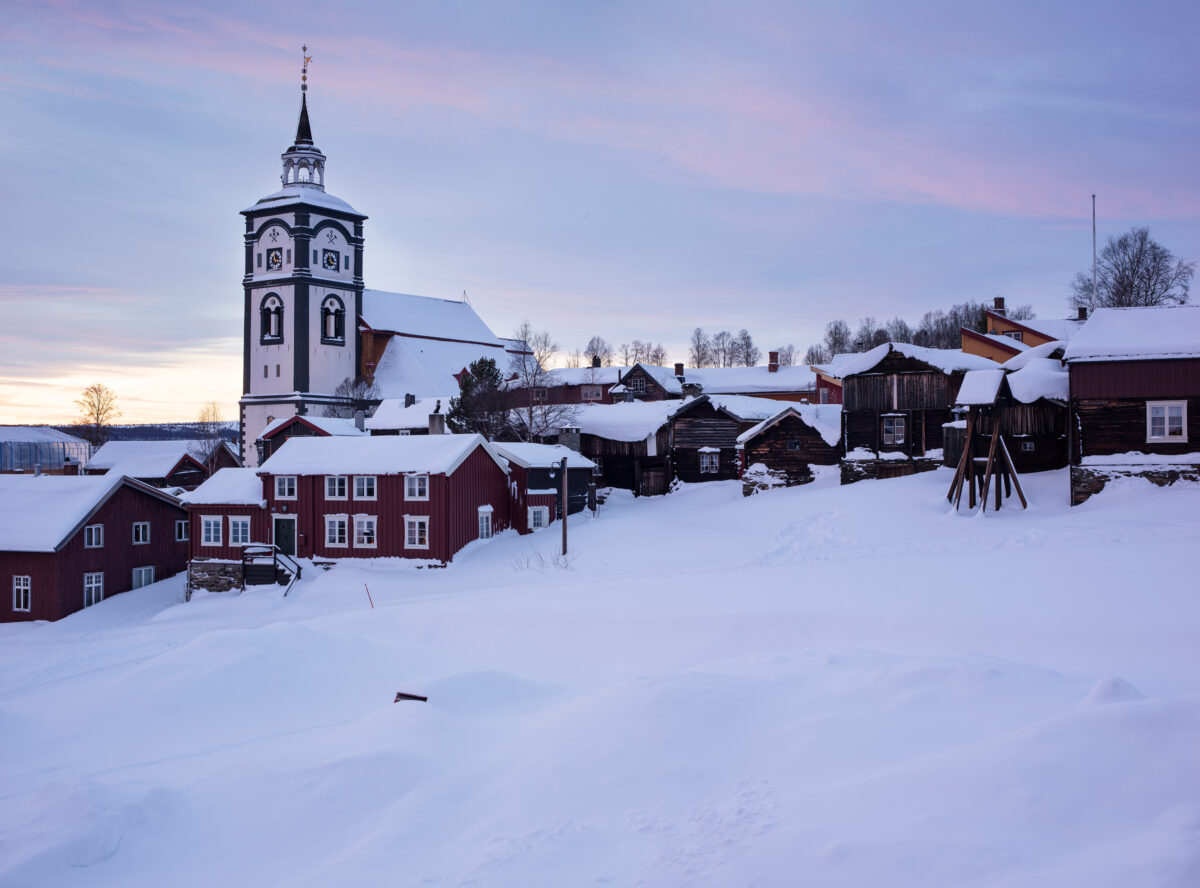 En snødekt landsby med trehus og en stor kirke med klokketårn ved soloppgang eller solnedgang.