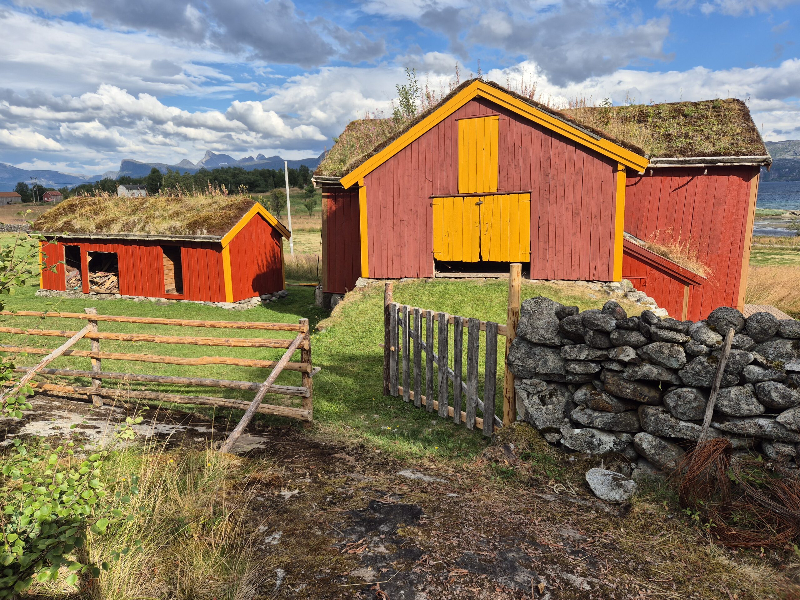 Tre røde trelåver med gule dører og gresstak står nær et steingjerde og en treport i et landlig landskap under en delvis overskyet himmel.