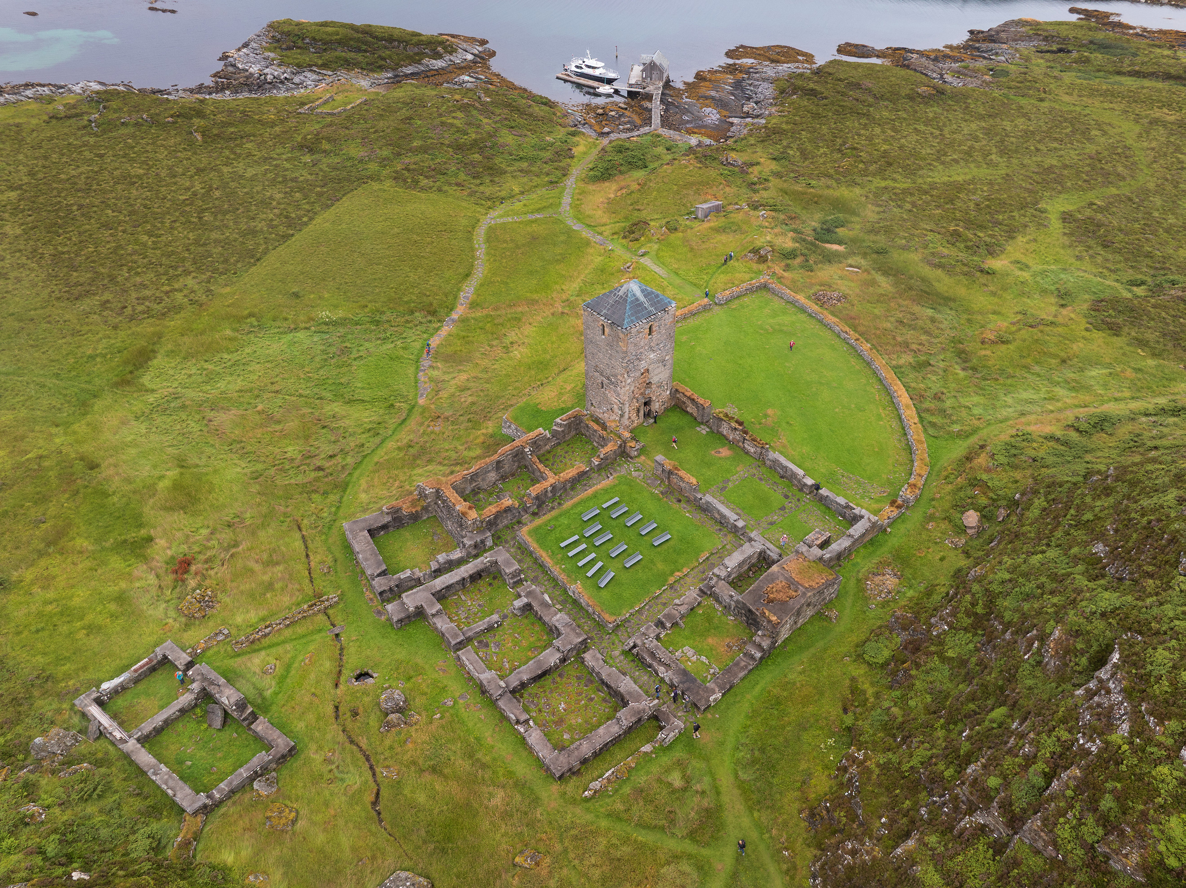 Flyfoto av en borgruin av stein med et sentralt tårn, gresslandskap rundt og en brygge med en båt ved vannet i bakgrunnen.