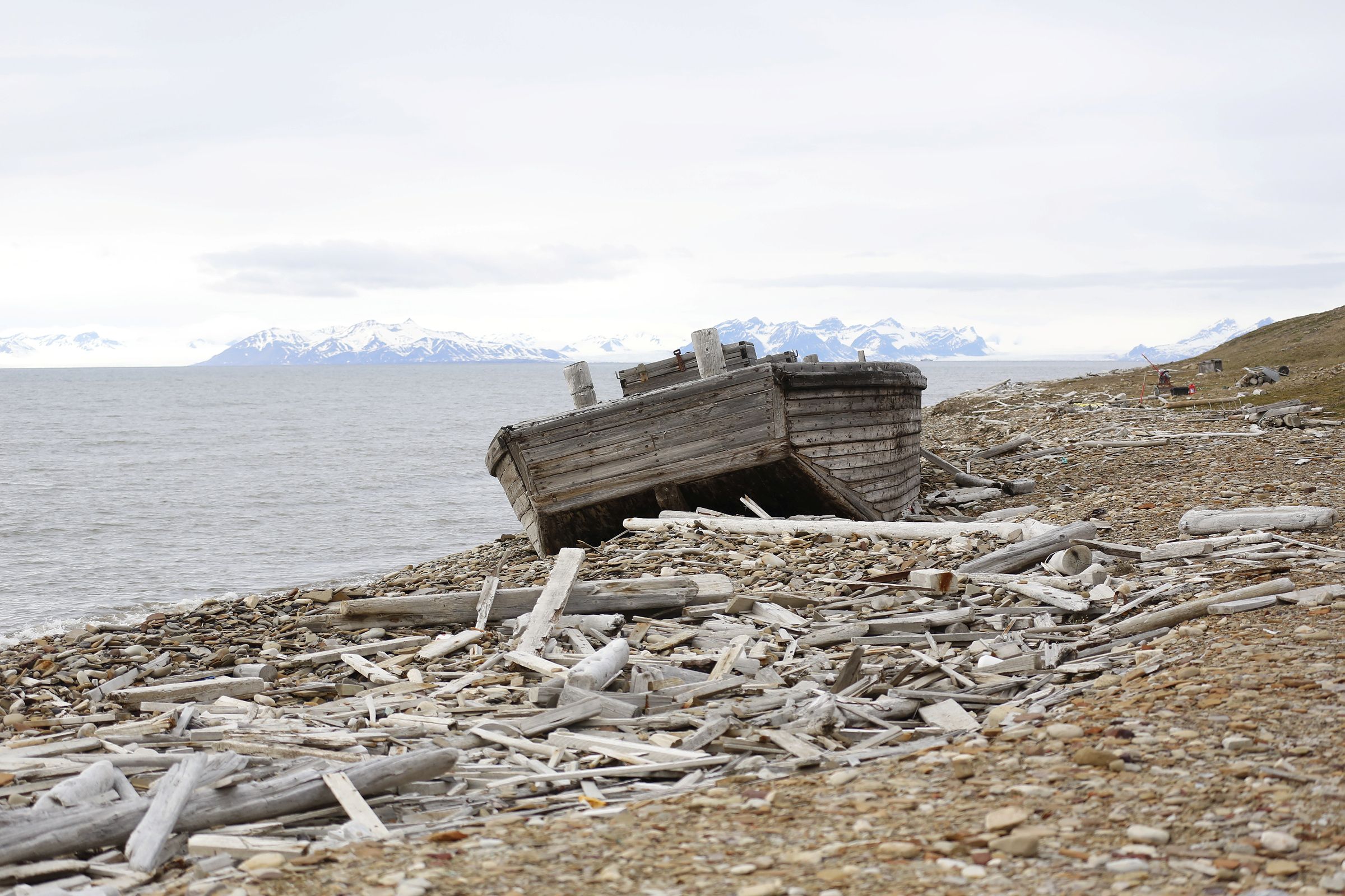 En gammel trebåt hviler på en steinete, drivvedkledd strand med snødekte fjell i det fjerne, på den andre siden av vannet, under en skyet himmel.