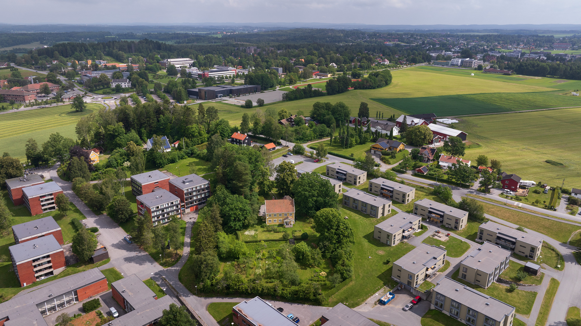 Flyfoto av et forstadsområde med boligblokker, hus, grønne trær og åpne jorder på en delvis overskyet dag.