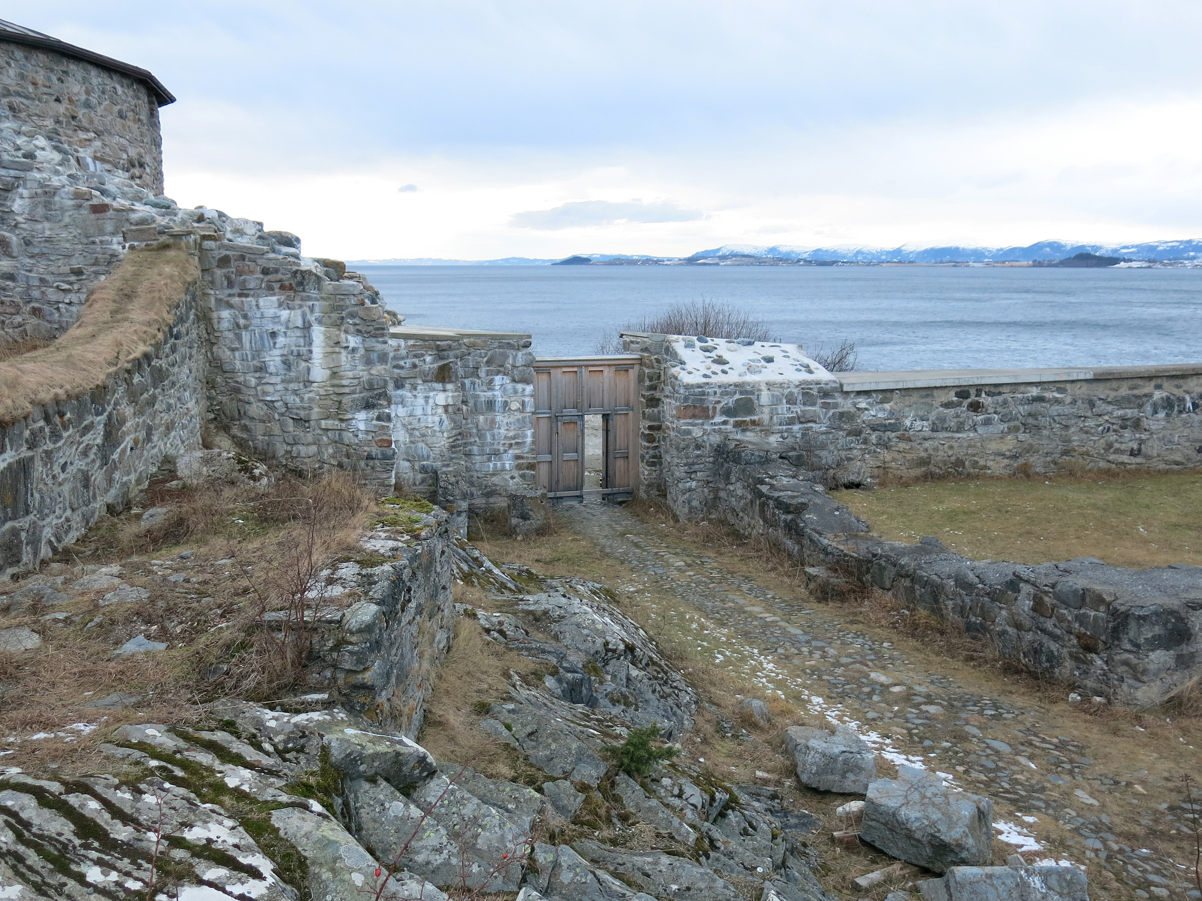 Steinmurer og en treport gir utsikt over et vann med snødekte fjell i det fjerne under en overskyet himmel.