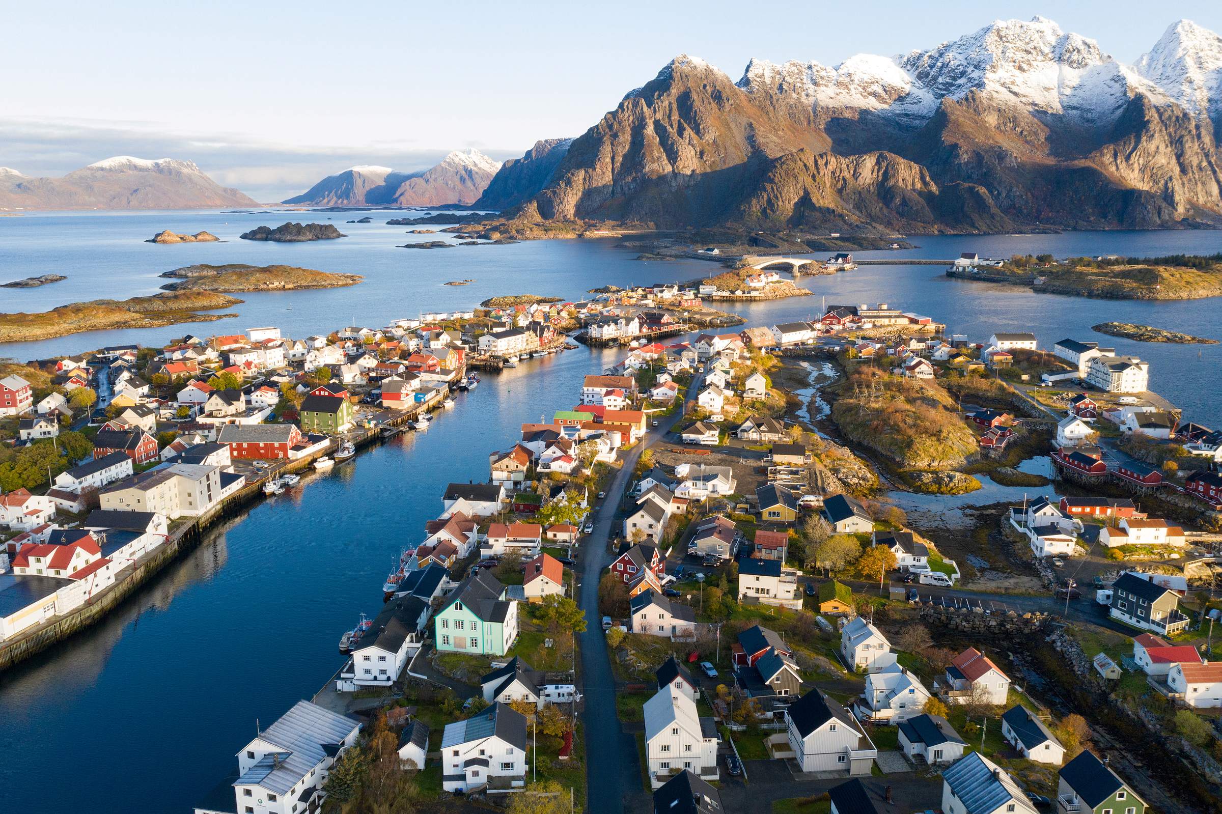 Luftfoto av en kystlandsby med fargerike hus, omgitt av vann og snødekte fjell under klar himmel.