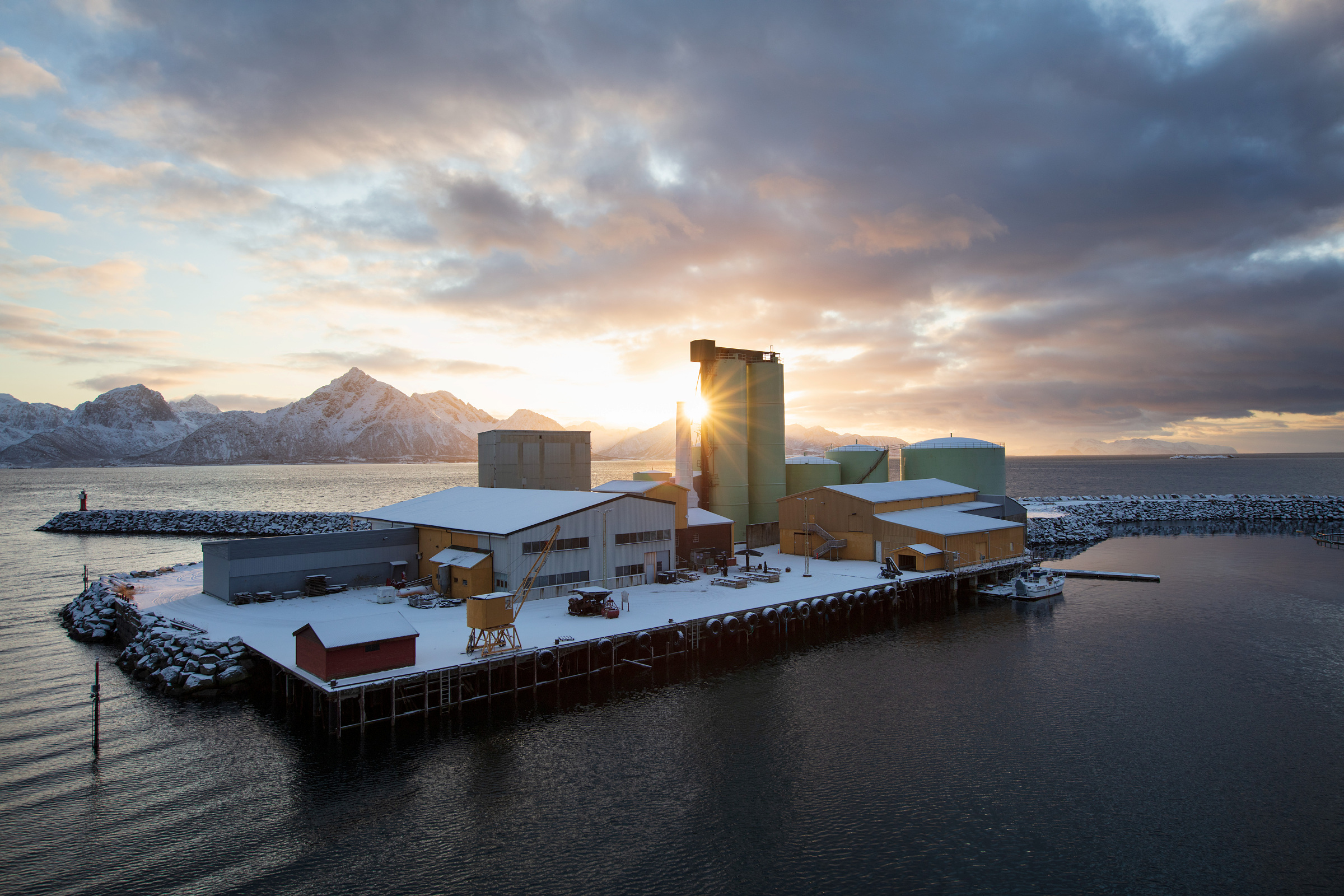 Riksantikvaren lyser ut 3,4 millionar kroner til bygder, byar, næring og industri 1 Industrianlegg på en snødekt brygge som strekker seg ut i vannet, med fjell i bakgrunnen og solnedgang bak bygningene.