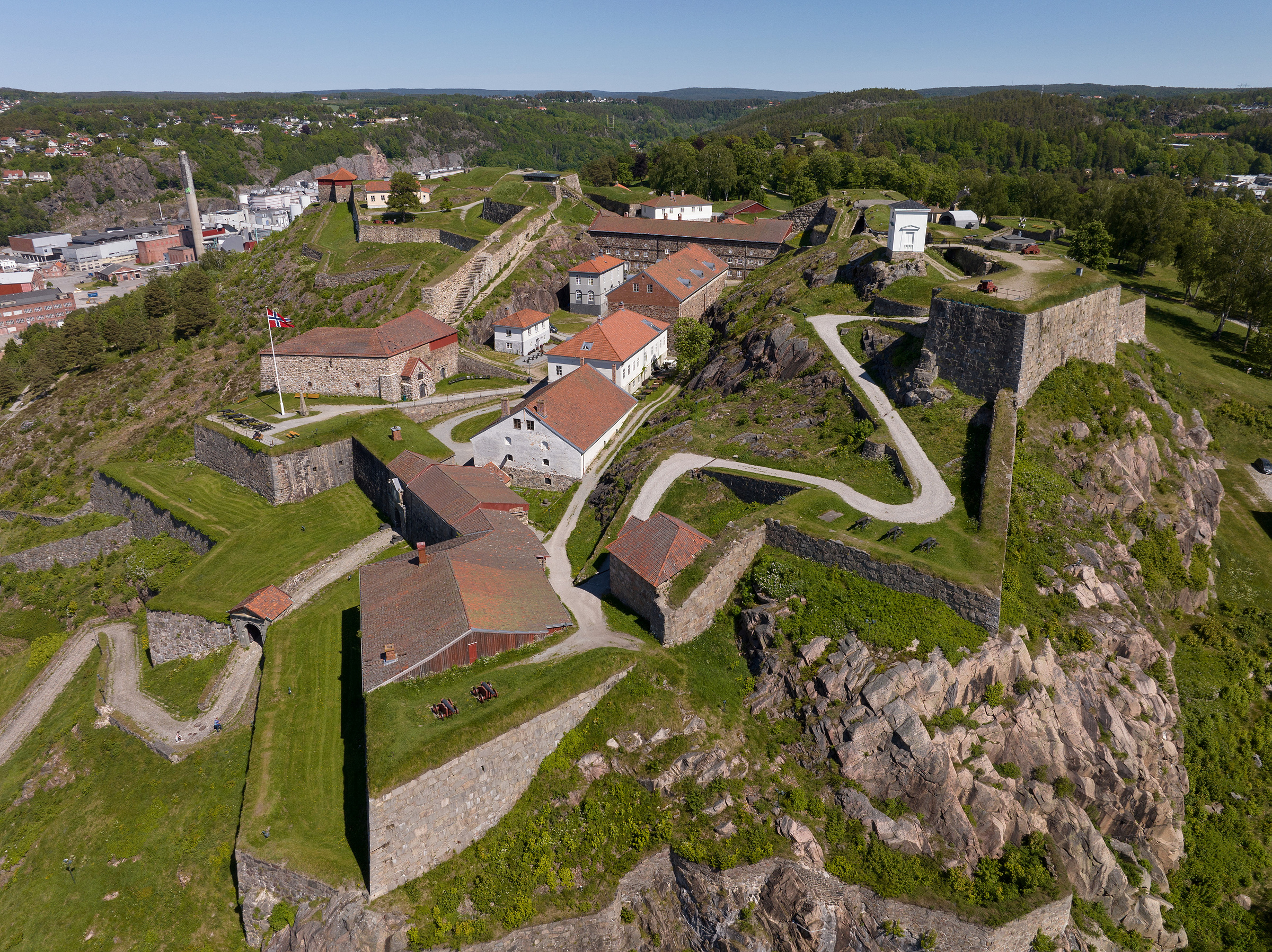 ICOMOS Norges fagdag: (U)FRED. Kulturarv – våpen eller bro? 2 Dronefoto av Fredriksten festning i Halden. Festningen vises i sollys, på bakketoppen sin, med steinmurer, bygninger med røde tak, svingete stier og grøntområder rundt.