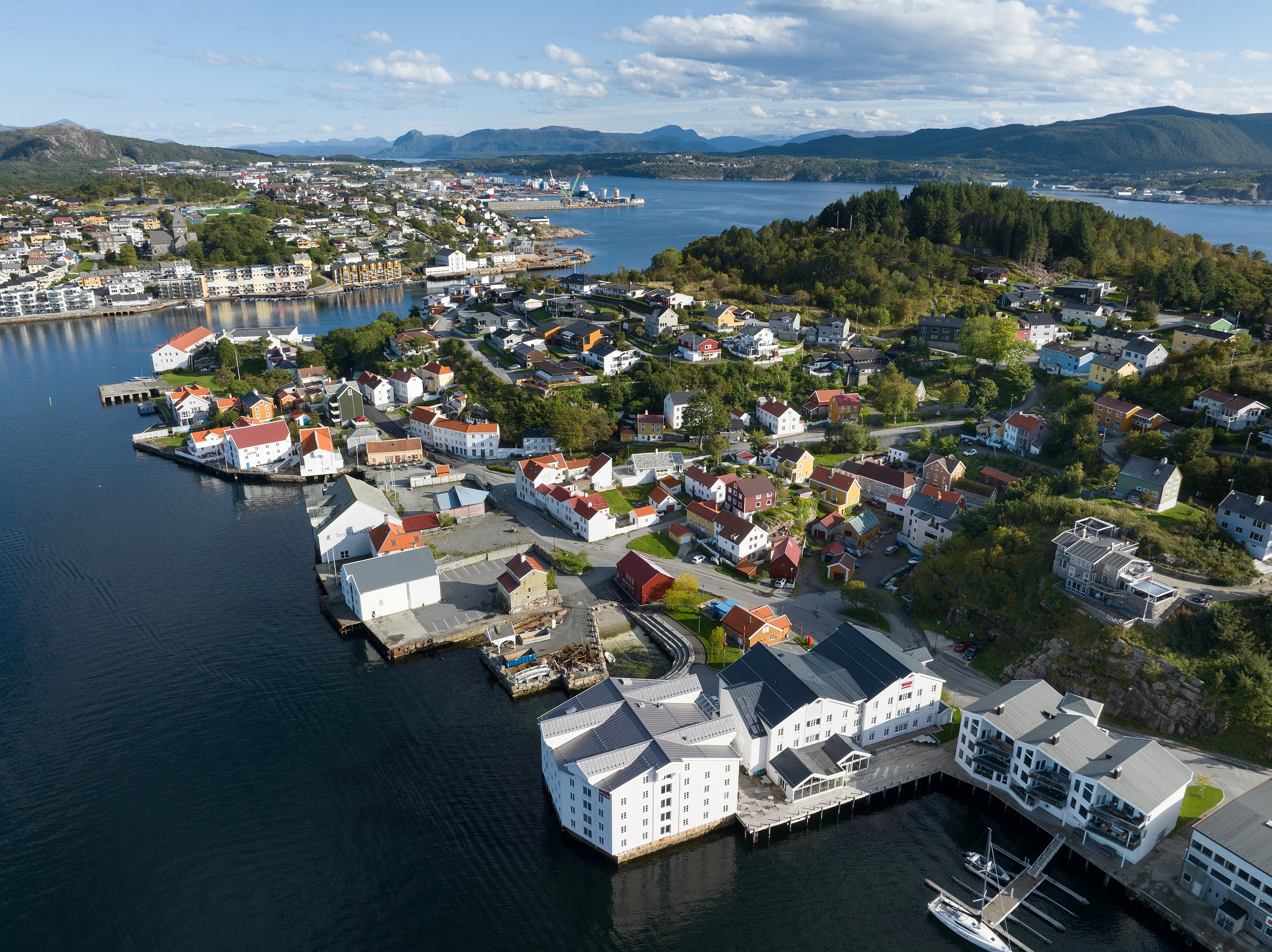 Flyfoto av en kystby med fargerike hus, brygger og båter, omgitt av vann og grønne åser under en delvis overskyet himmel.