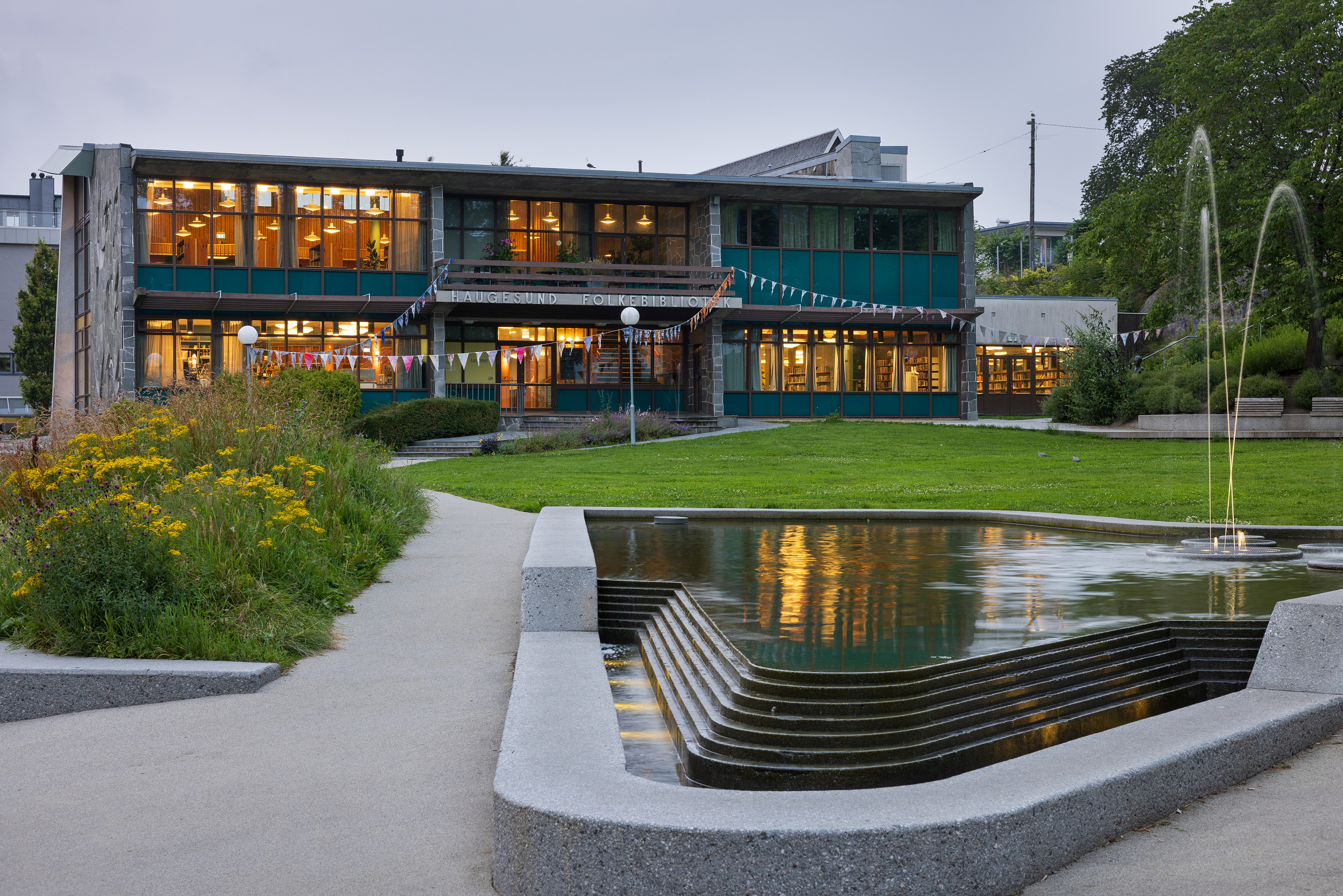Moderne toetasjes bygning med store vinduer, fargerike vimpler og et "Der vi møtest"-skilt over inngangen, som grenser til et grøntområde med geometrisk vannspeil og fontene.