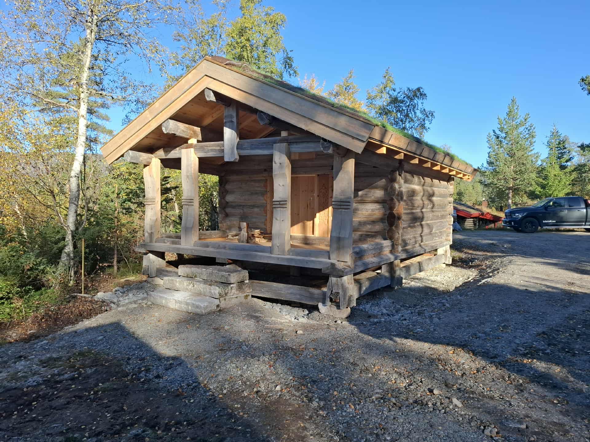 Et lite stabbur med overbygd veranda står på en gruslagt lysning, omgitt av trær og en parkert svart bil i bakgrunnen.