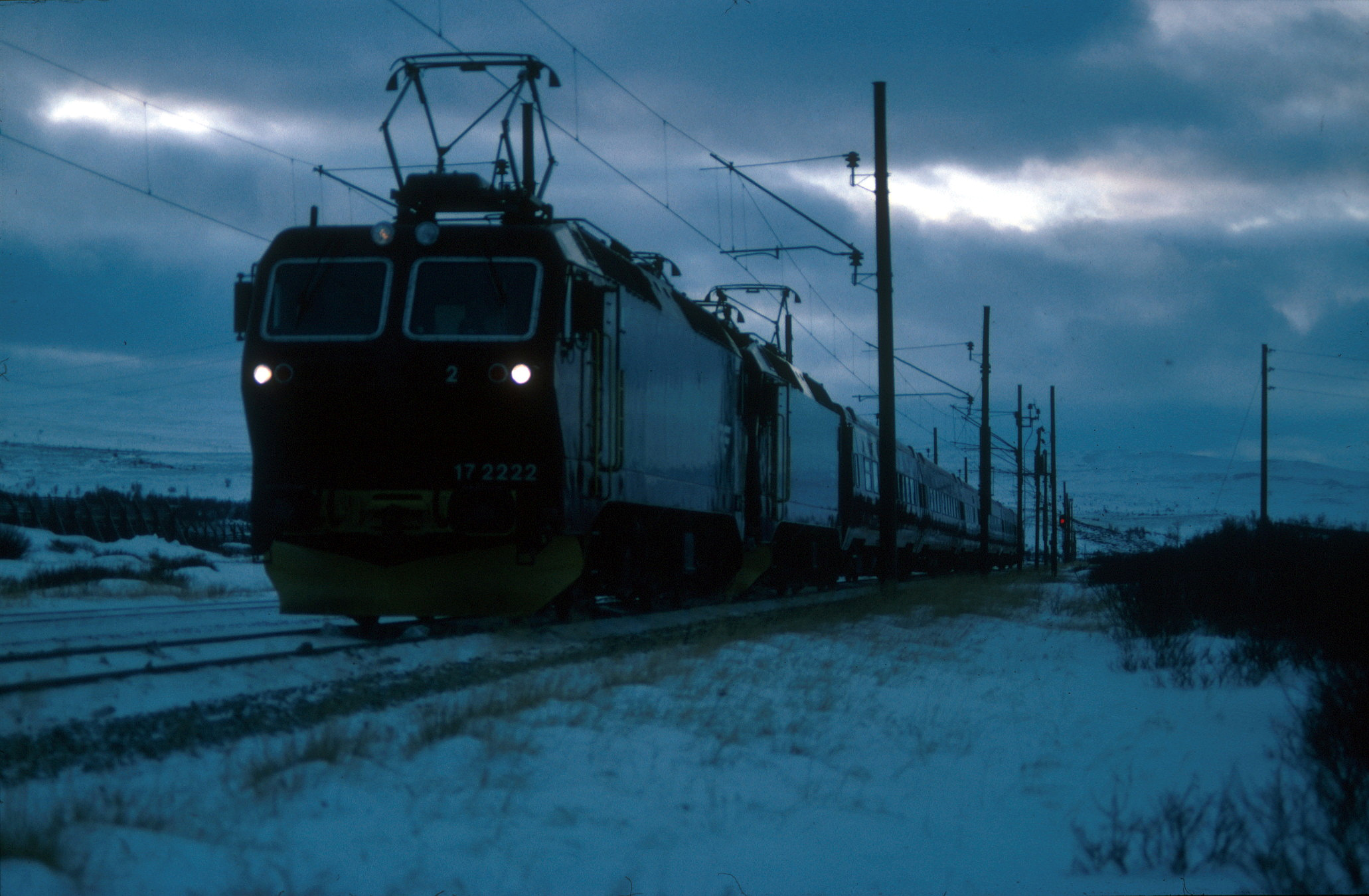 Et mørkt, elektrisk tog kjører gjennom et snødekt landskap under en overskyet himmel, med kraftledninger langs skinnene.