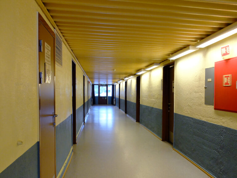 En lang, tom gang med gult tak, beige og blå vegger, lukkede brune dører og lysstoffrør i taket. På høyre vegg er det montert brannsikkerhetsutstyr.