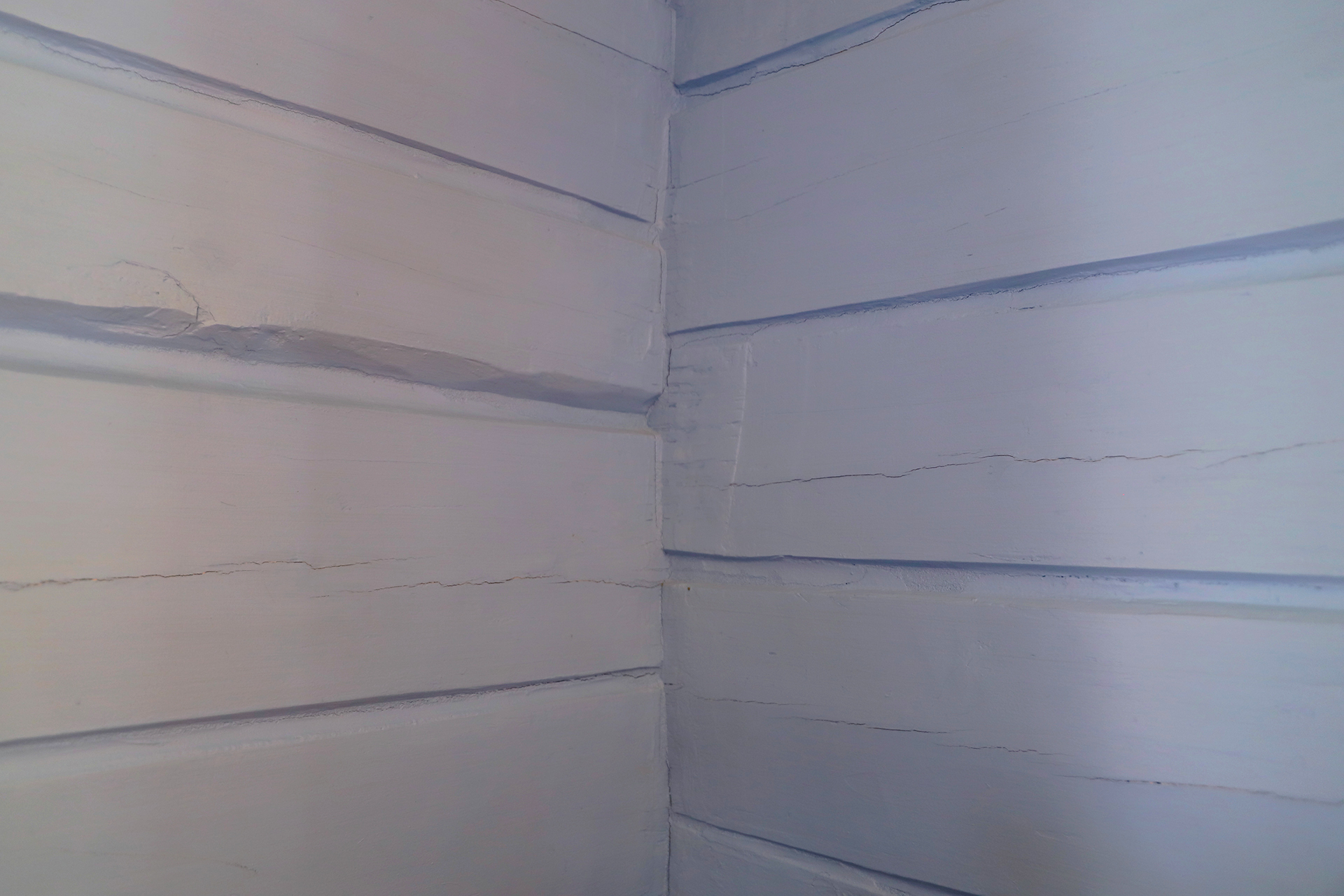 Hjørnebilde av to kryssende trevegger med synlige horisontale planker og tydelige sprekker i malingen.