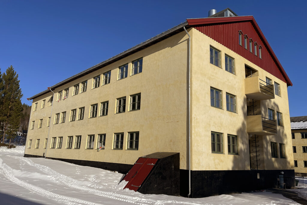 En gul bygning i tre etasjer med flere vinduer, rødt saltak og en liten metalldør i kjelleren, omgitt av snø under en klarblå himmel.