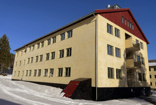 En gul bygning i tre etasjer med flere vinduer, rødt saltak og en liten metalldør i kjelleren, omgitt av snø under en klarblå himmel.