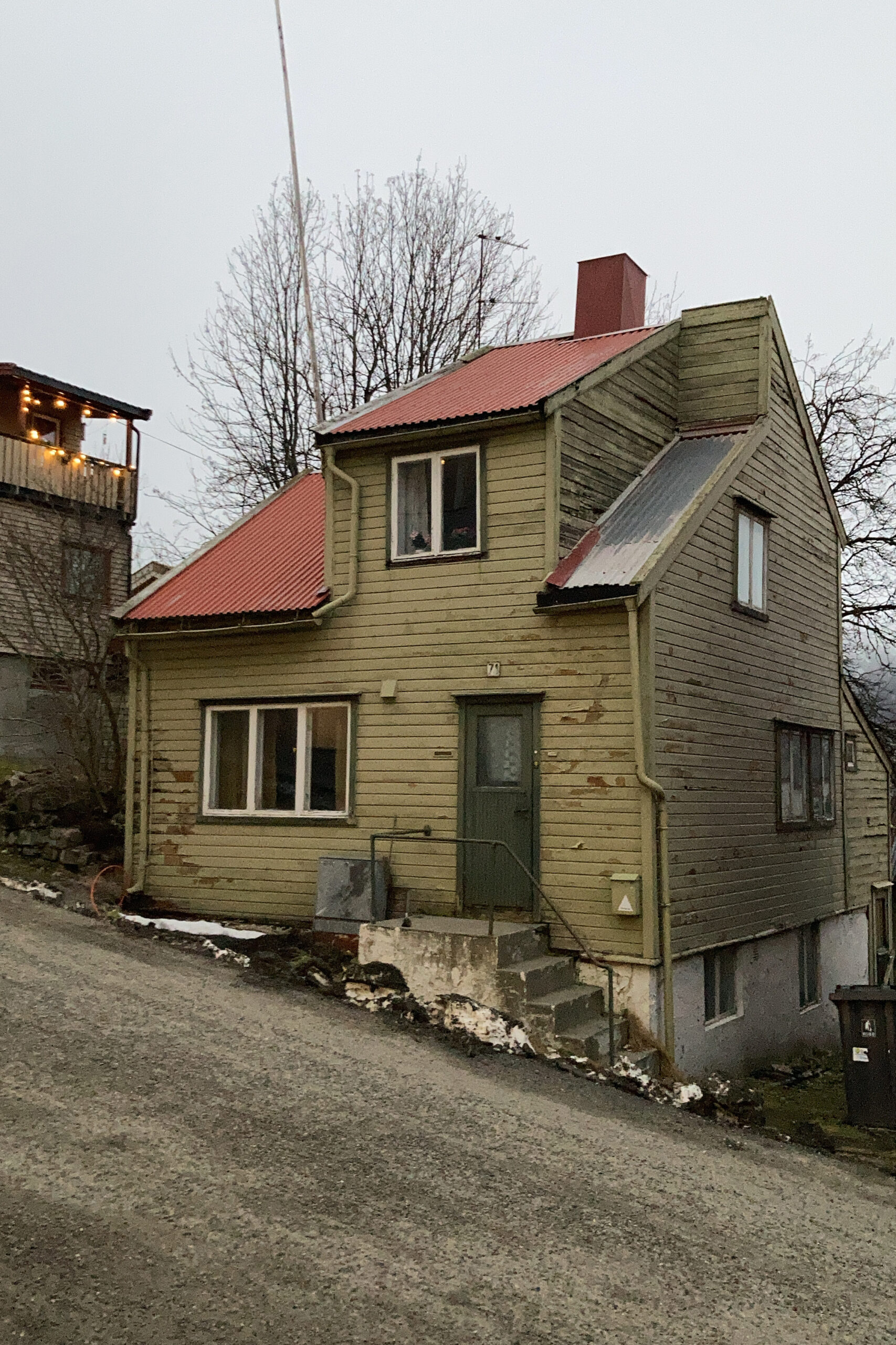 Et toetasjes trehus med rødt tak, grønn dør og forvitret eksteriør ligger i en skrånende gate med nakne trær i bakgrunnen.