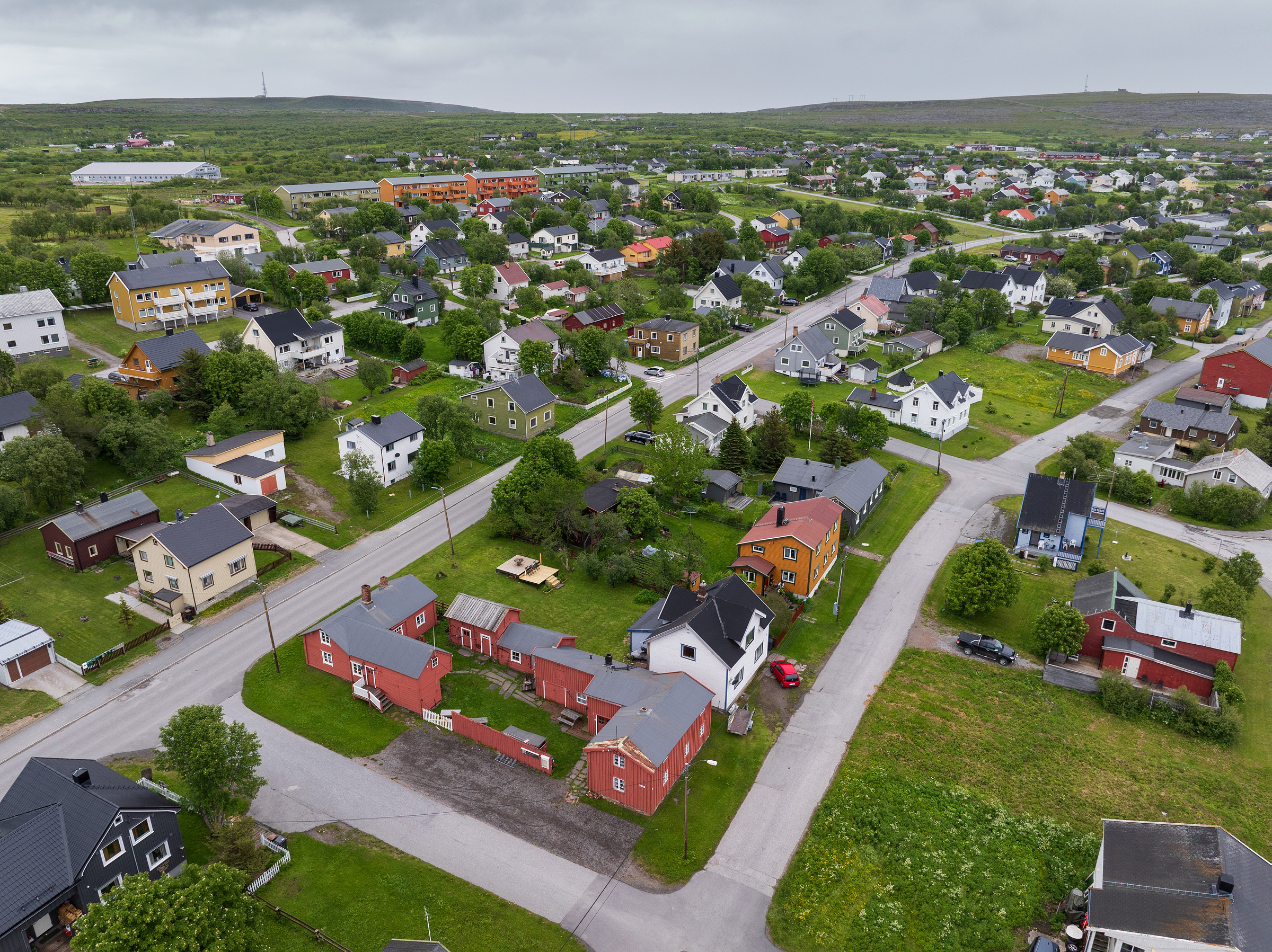 Flyfoto av en liten by med fargerike hus, gater omkranset av trær og grønne områder omkranset av åser under en overskyet himmel.