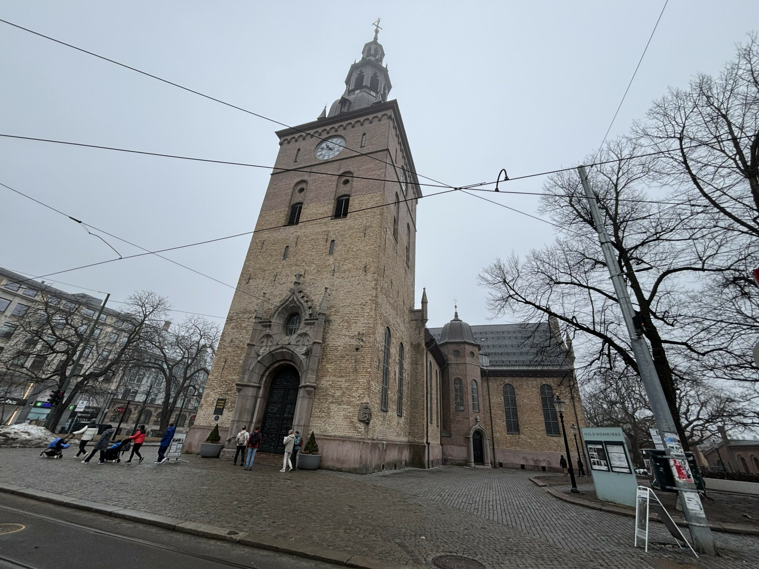 En stor steinkirke med klokketårn står på en brosteinsbelagt gate; folk spaserer i nærheten, og løvløse trær omkranser bygningen på en overskyet dag.