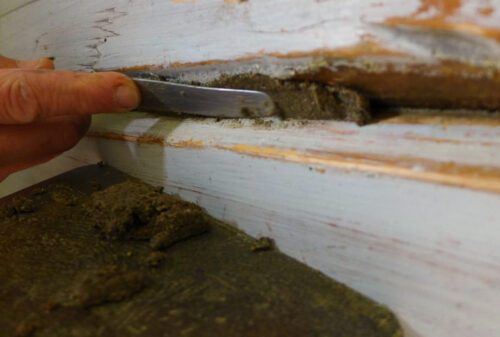 En hånd bruker et metallverktøy til å skrape eller fjerne en brun, kittlignende substans fra en sprekk i en malt treflate.