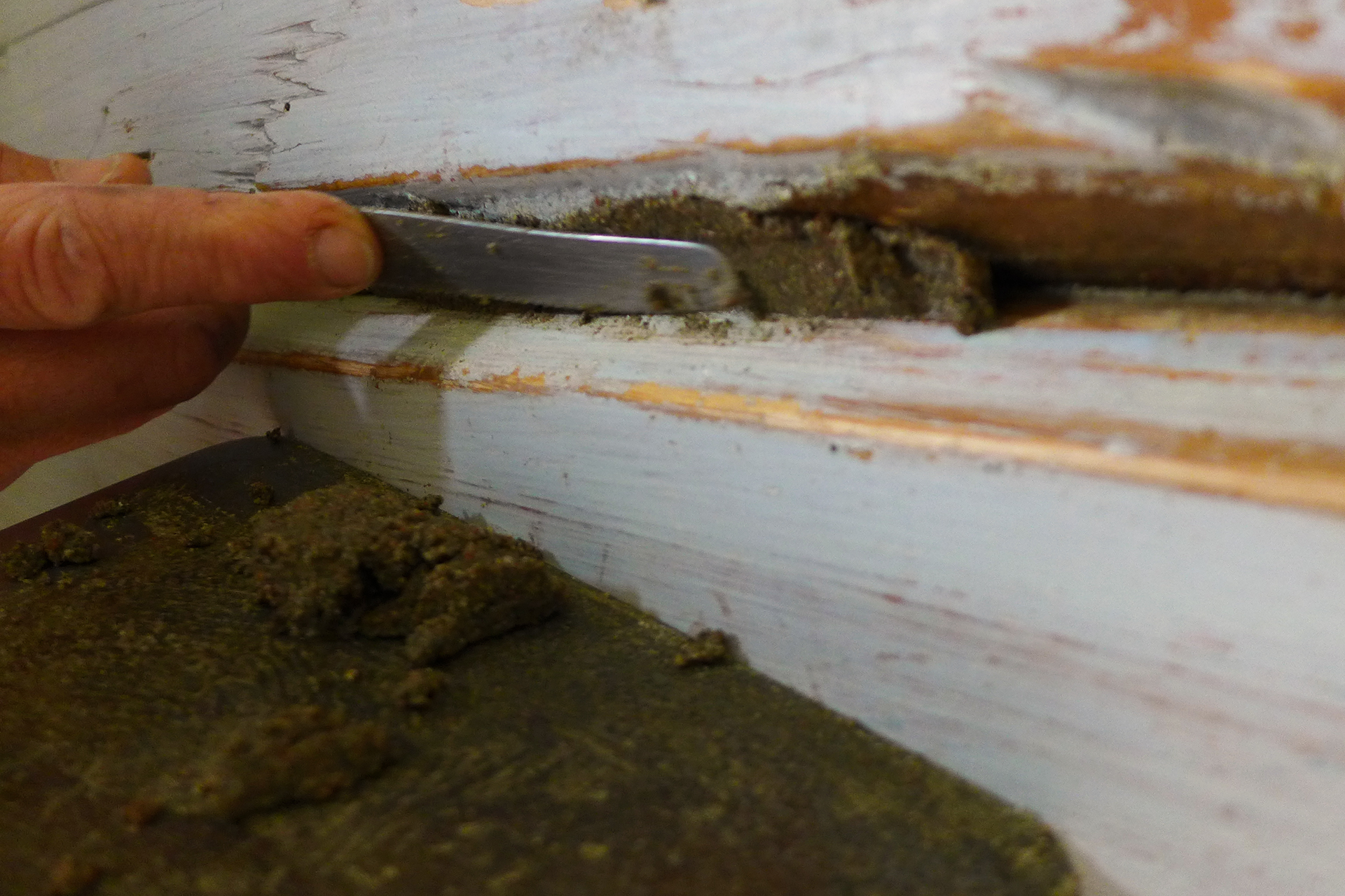 En hånd bruker en sparkelkniv til å fjerne gammel, inntørket kitt eller sparkelmasse fra en sprekk i en treflate eller vindusramme.